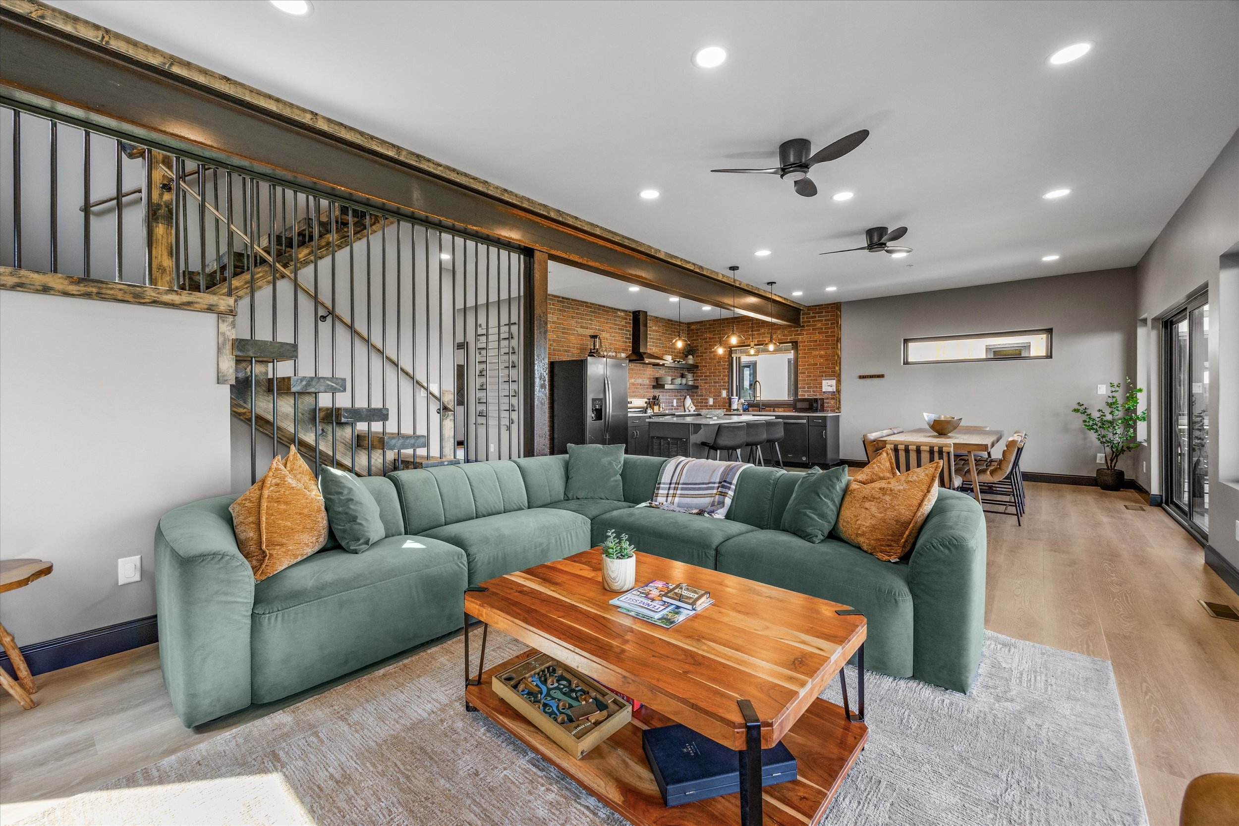 Open-concept living room and kitchen with brick accent wall, black kitchen cabinets, bar stools, green couch with throw pillows, wooden coffee table with magazines, and ceiling fans.