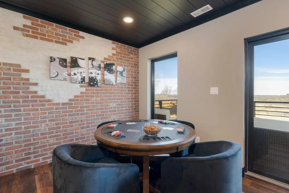 A round poker table with chips and cards in a cozy room with brick and white walls, large windows showing a clear, sunny day outside, and dark ceiling.