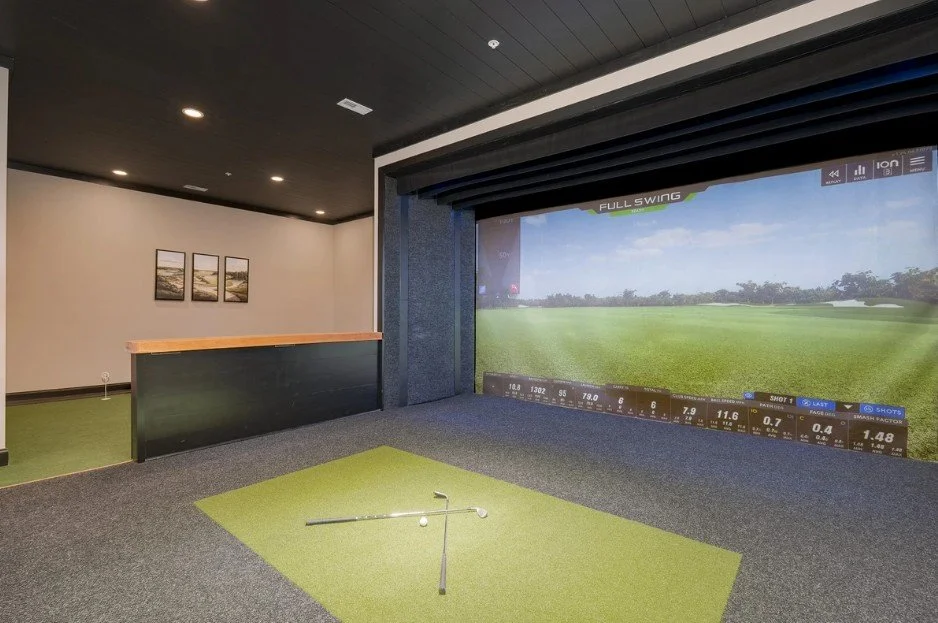 Indoor golf simulator room with a large screen showing a golf course, a golf club, and a golf ball on a green mat.