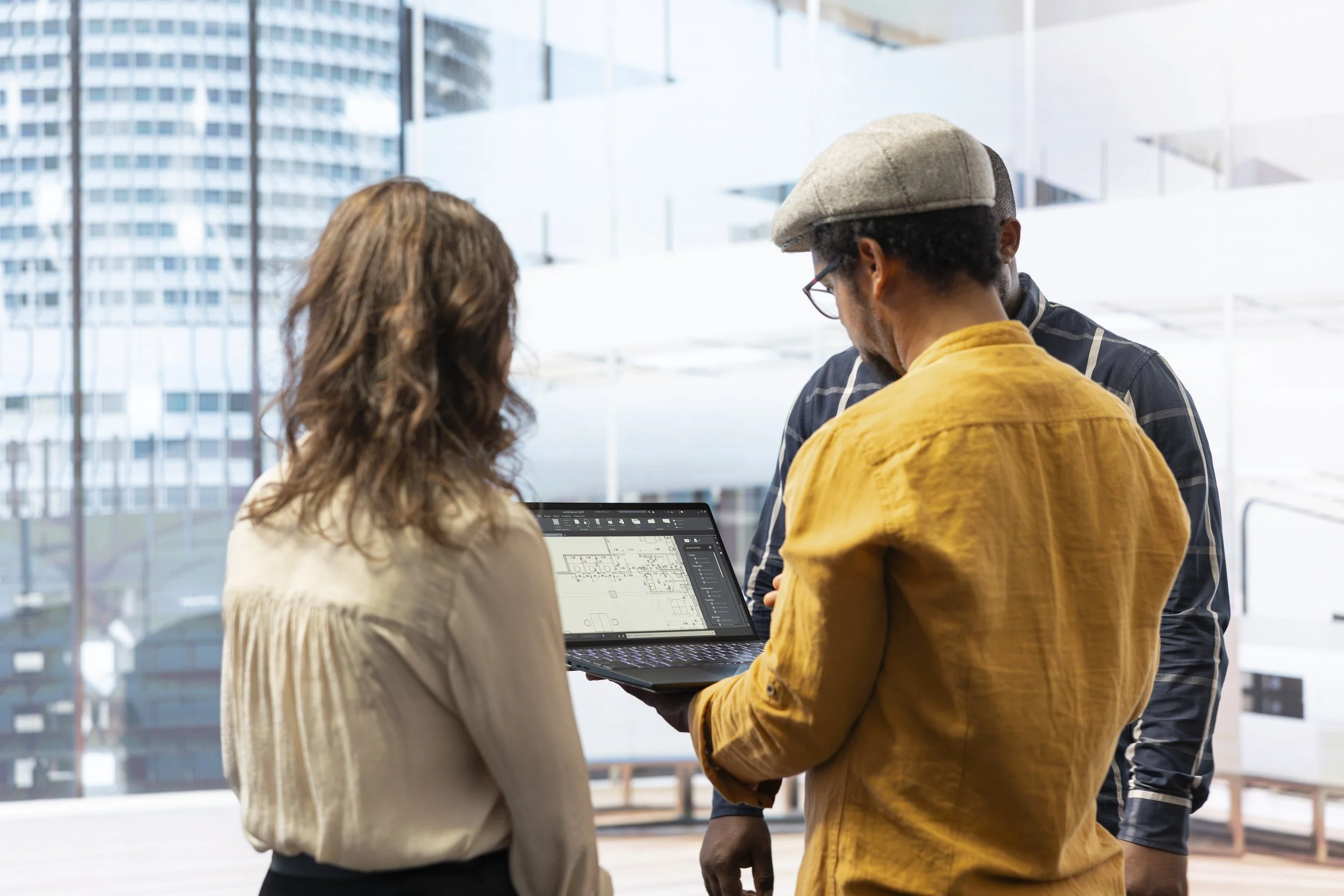 Three people in discussion during a meeting with a laptop displaying architectural plans, in a bright office with large windows showing a cityscape.