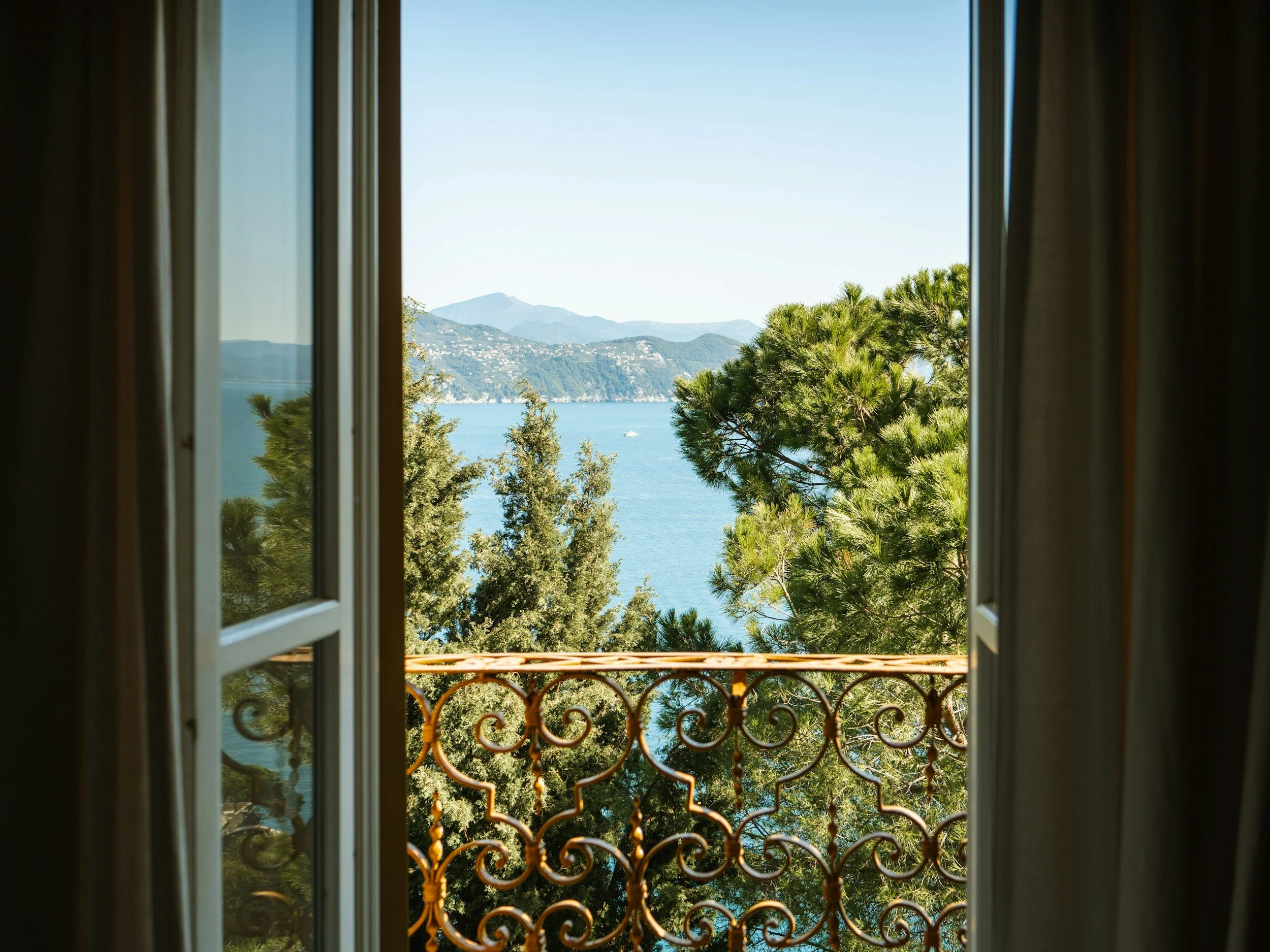 View of distant mountains, trees, and a body of water seen through an open window with a decorative metal balcony railing.