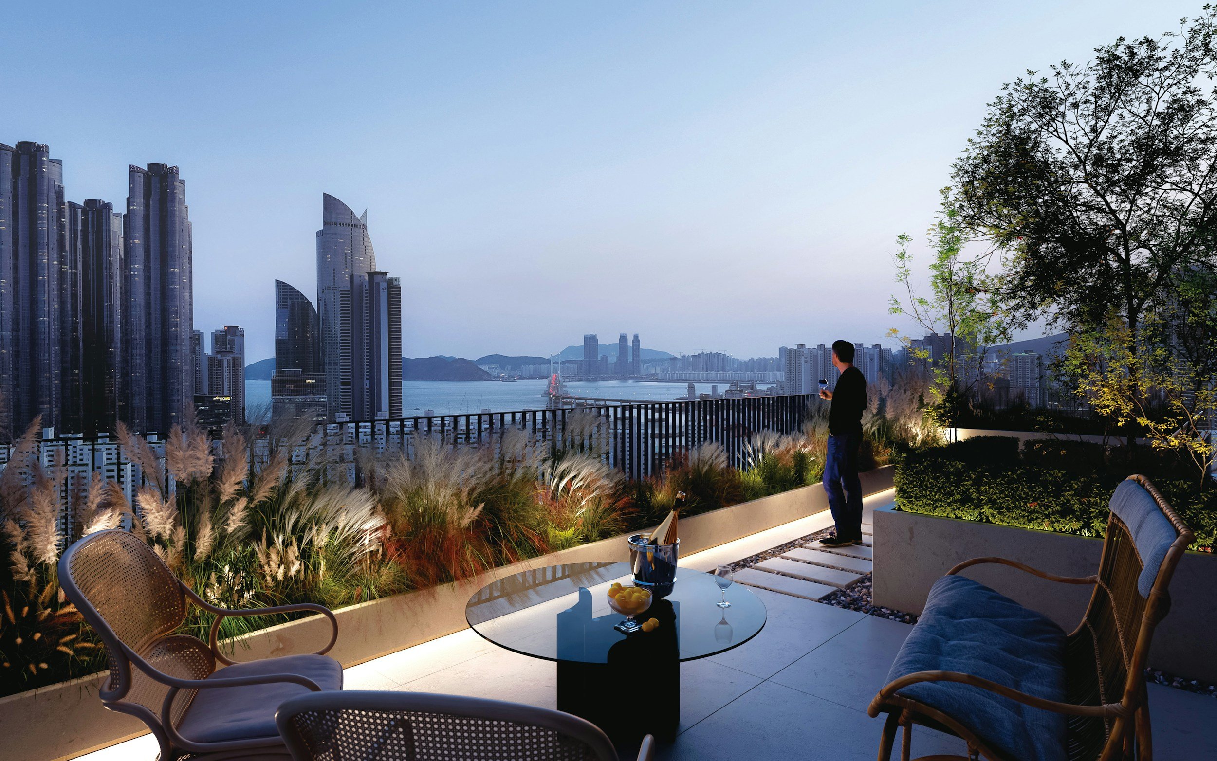 A rooftop balcony with outdoor furniture, plants, and a view of a city skyline with high-rise buildings and a body of water in the distance.