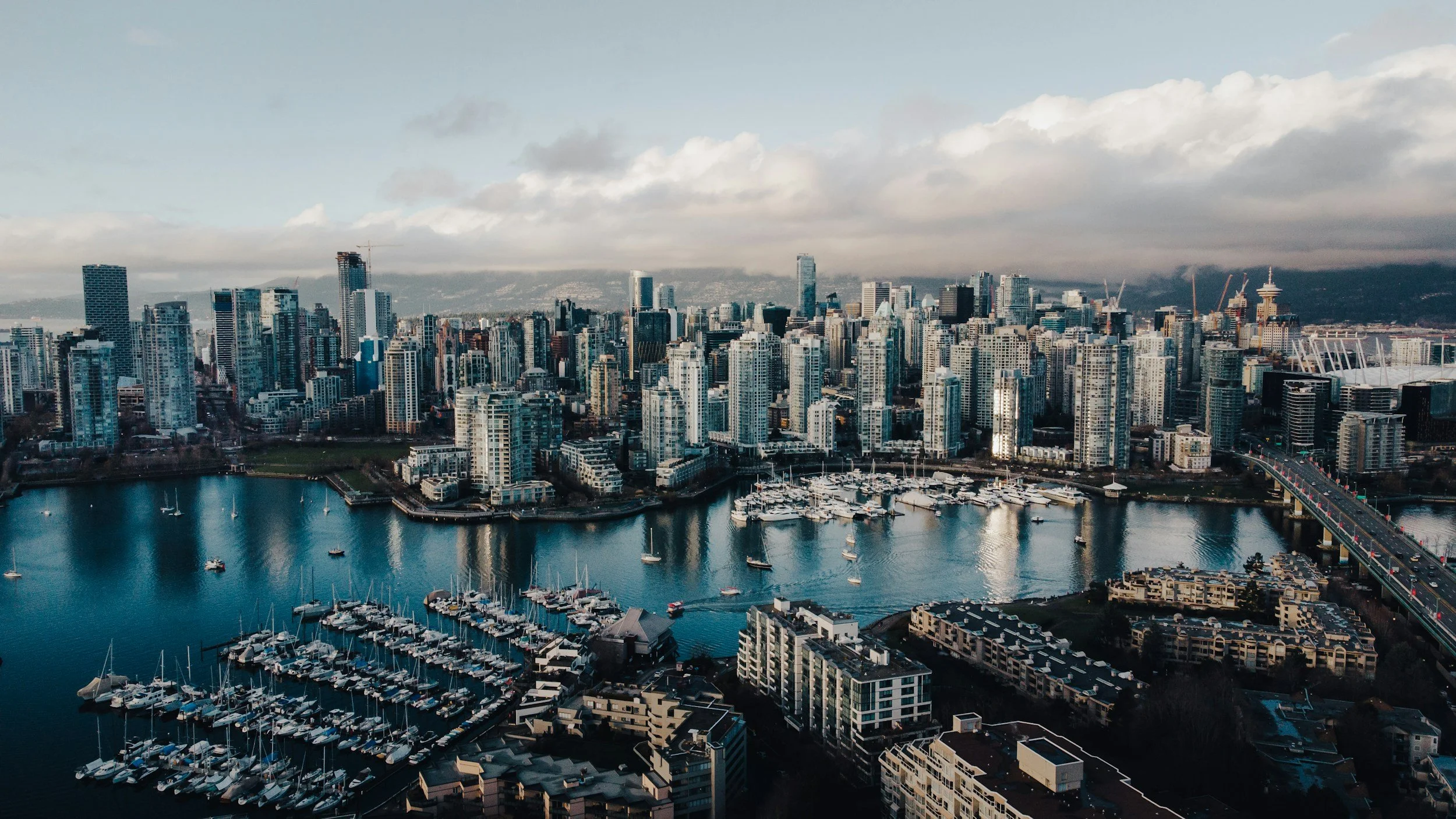 A city skyline with numerous high-rise buildings and skyscrapers, a body of water with boats docked at marinas, and a bridge over the water. Clouds are visible in the sky.