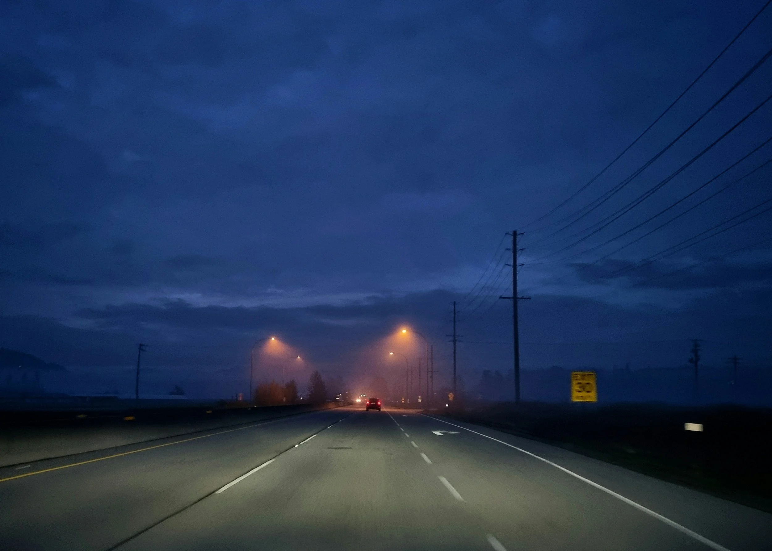 A dark highway at night with a few cars, streetlights, and power lines visible against a cloudy, blue sky.
