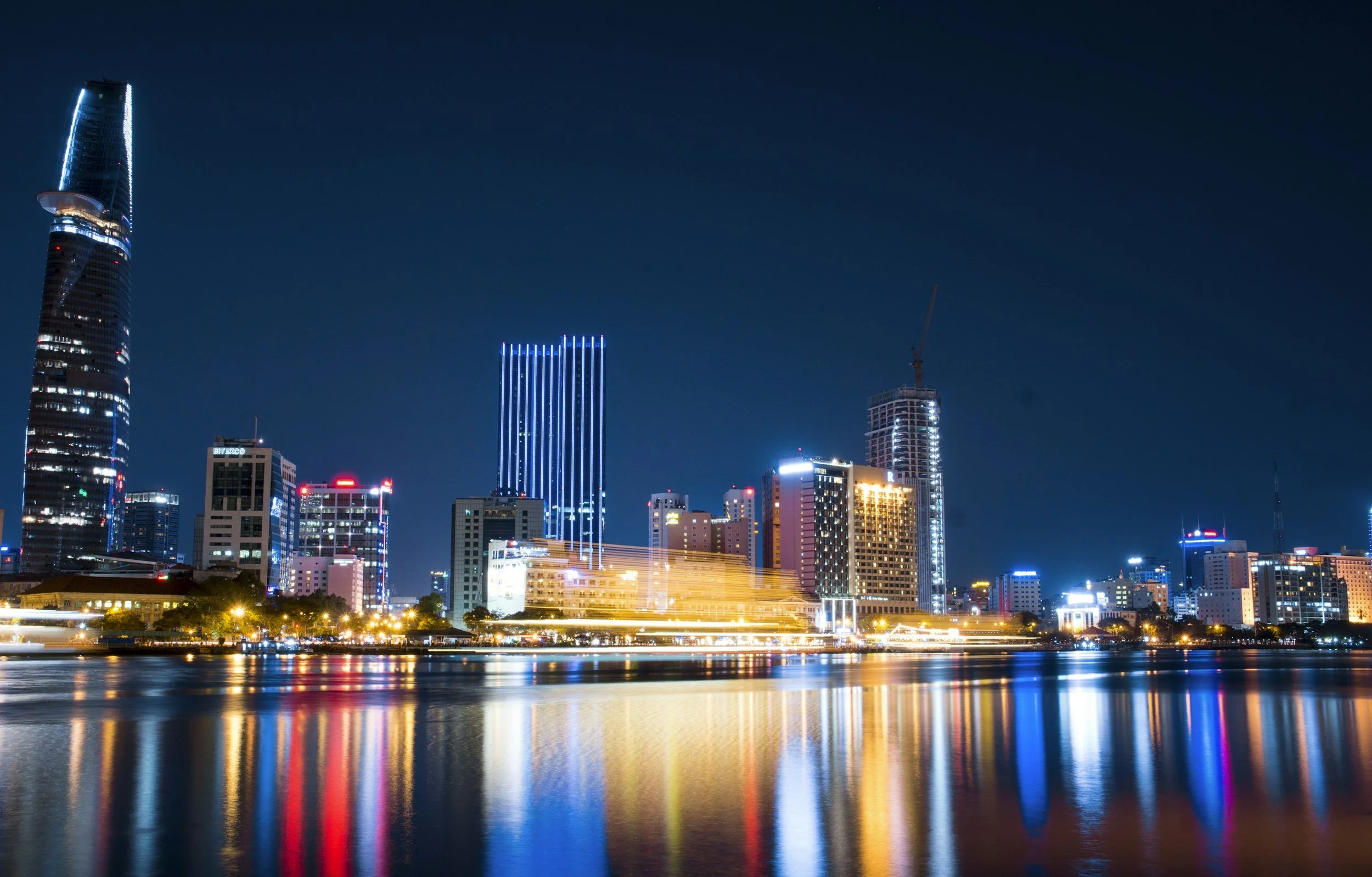 Night view of a city skyline with illuminated buildings reflecting on a river, featuring tall skyscrapers with blue and white lights.