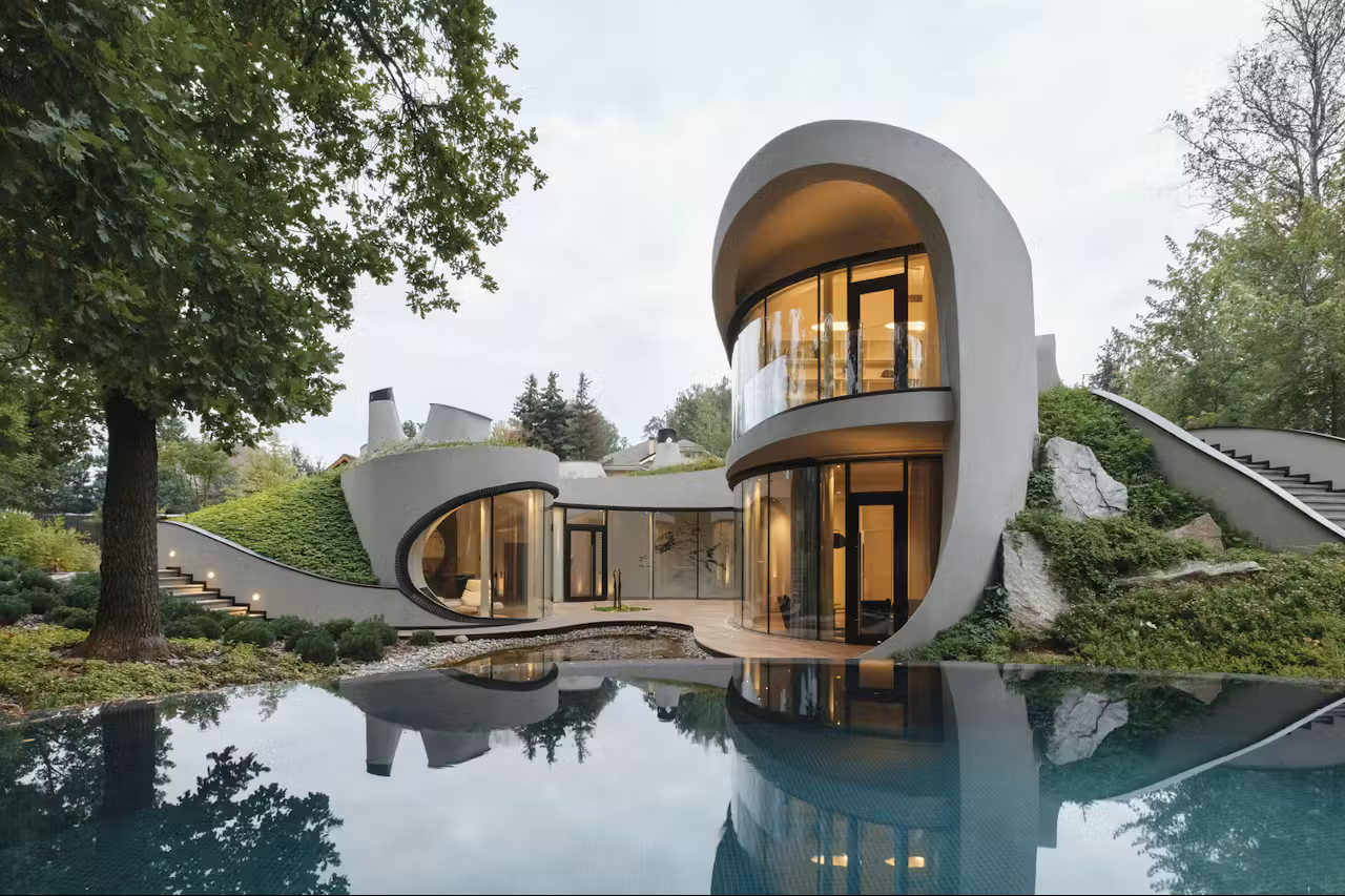 Modern house with curved architecture and large glass windows, surrounded by trees, with a reflecting pool in the foreground.