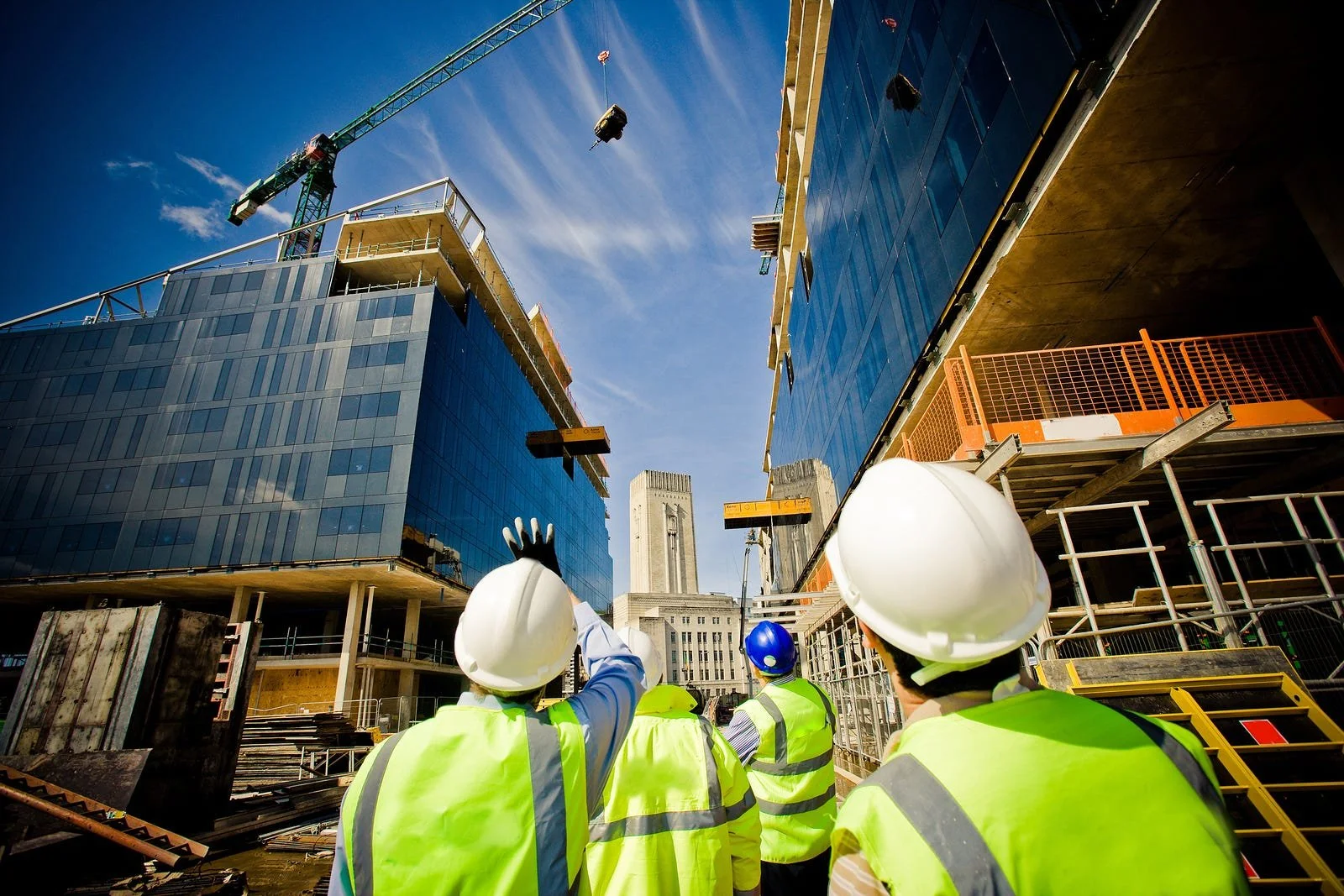 Construction workers wearing safety helmets and vests look up at the building site where two tall buildings are being constructed with glass facades and a crane working overhead, with a historic-looking building in the background under a clear blue sky.