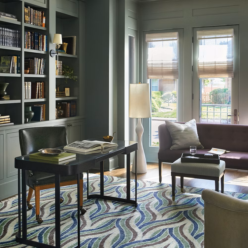 Living room with a black desk, a black leather chair, a purple sofa, a small striped ottoman, a white floor lamp, and large windows with beige blinds.