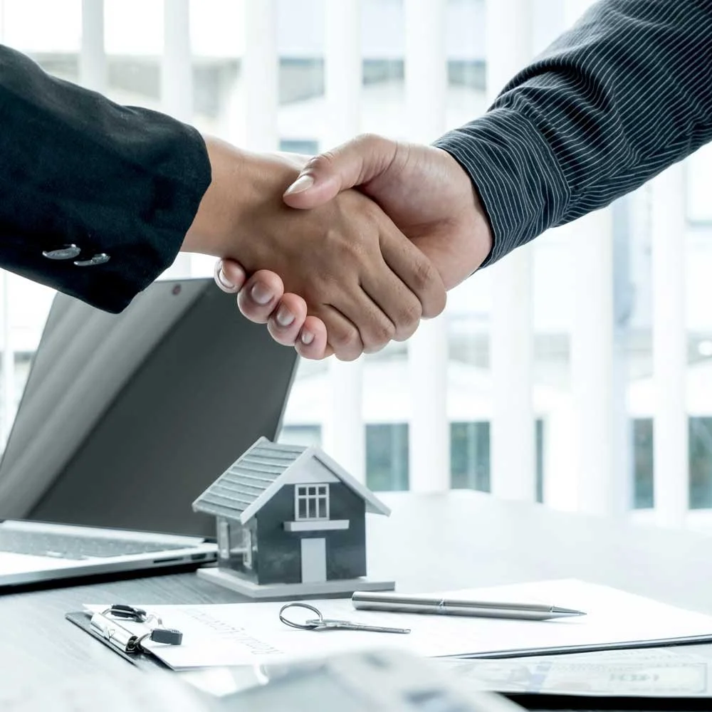 Two people shaking hands over a table with a small model house, keys, and a clipboard with documents, symbolizing a real estate agreement or deal.
