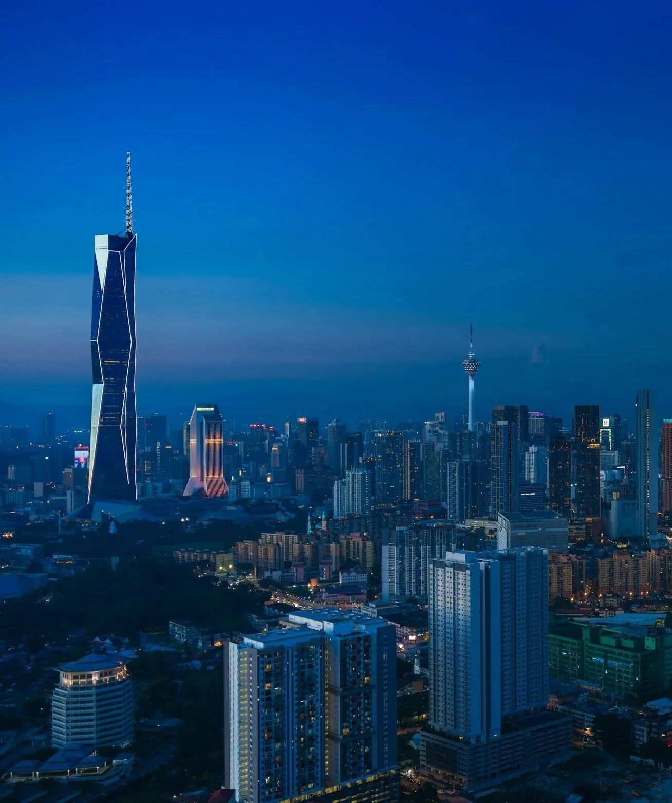 A city skyline at dusk with tall skyscrapers and a clear blue sky. Notable buildings include a modern glass skyscraper on the left and a tall tower with an observation deck towards the center-right.