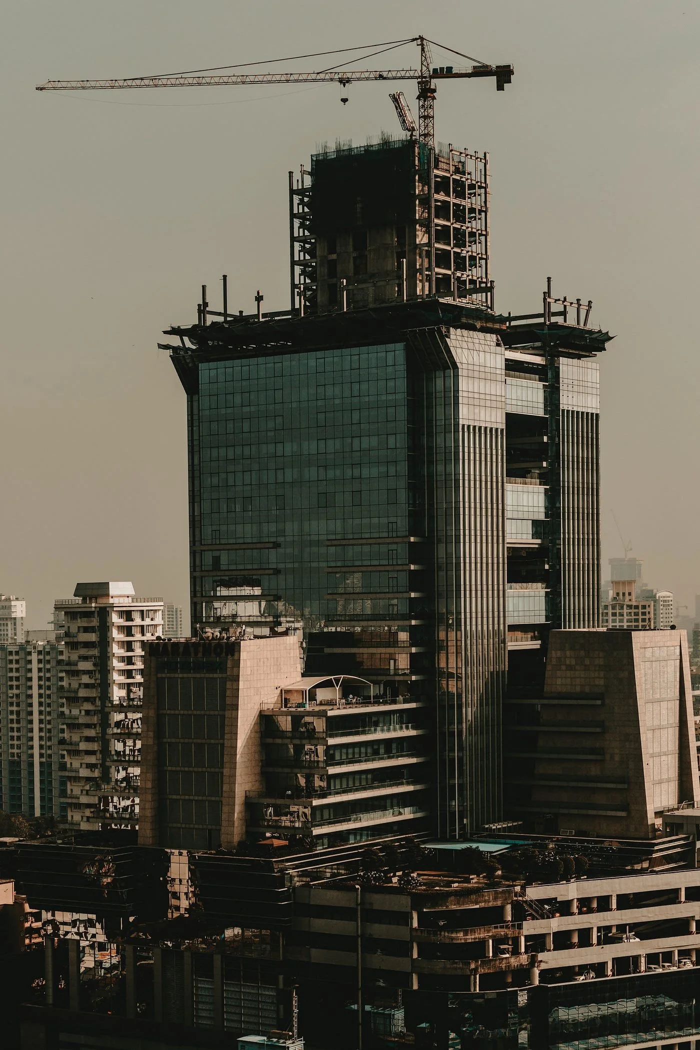 A tall modern skyscraper under construction with a crane at the top, surrounded by other high-rise buildings in an urban setting during sunset.