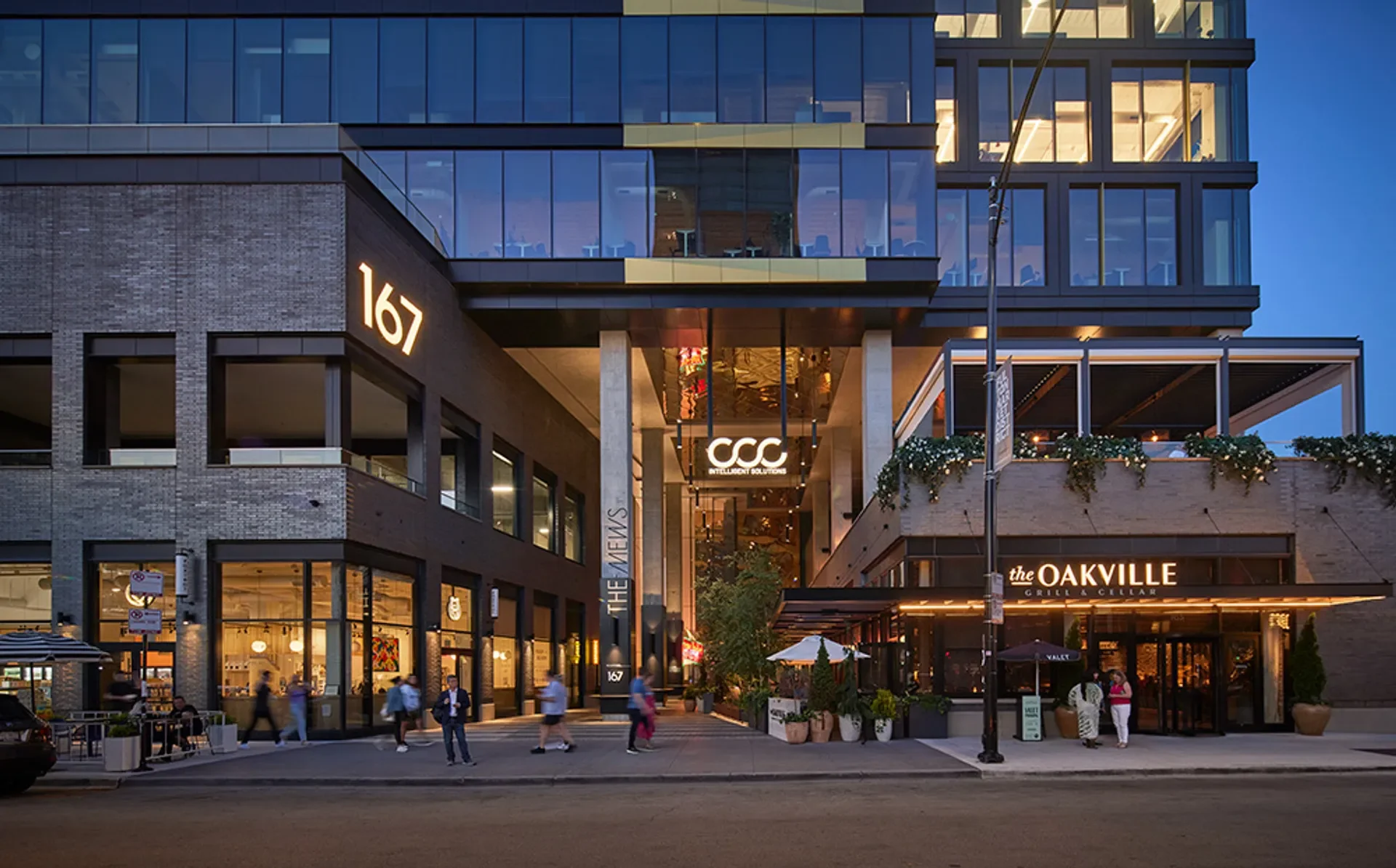 The exterior of a modern multi-story building with glass windows and signage for businesses including the Oakville Grill & Cellar, located at 167. The building features a mixture of brick and glass architecture, with outdoor seating and hanging lights visible outside during dusk.