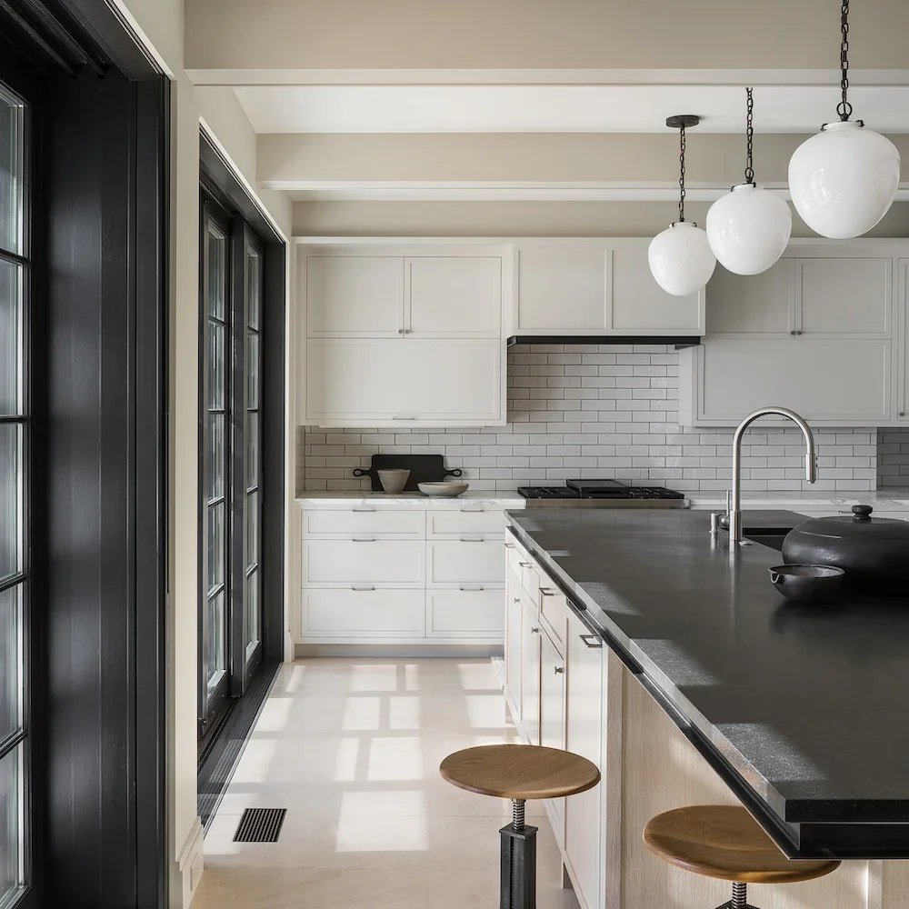 Modern kitchen with white cabinets, black countertop, white subway tile backsplash, pendant lights, and black-framed windows.