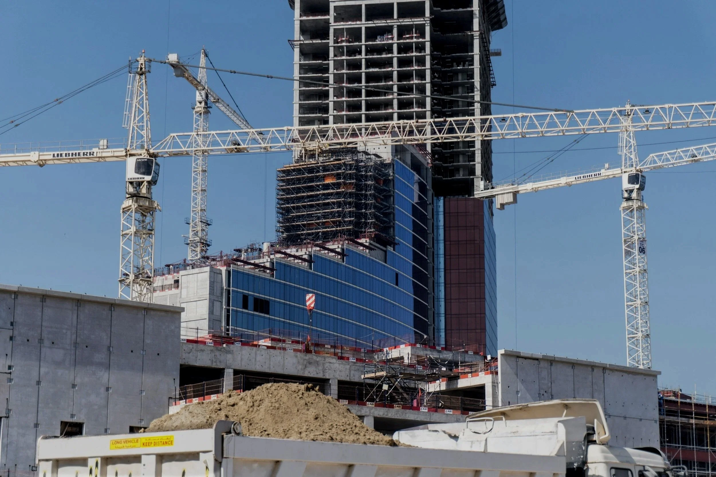 A high-rise building under construction with two large white cranes in the foreground, blue sky in the background, and a pile of dirt and construction barriers in the front.