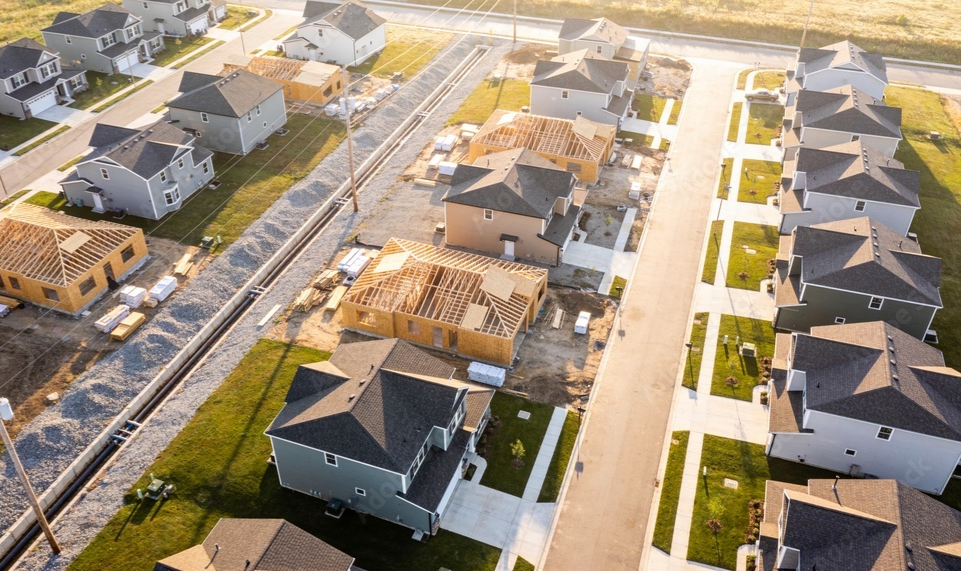 Aerial view of a neighborhood with houses under construction, some with roofs completed and others with framing still in progress, alongside paved sidewalks and green yards.