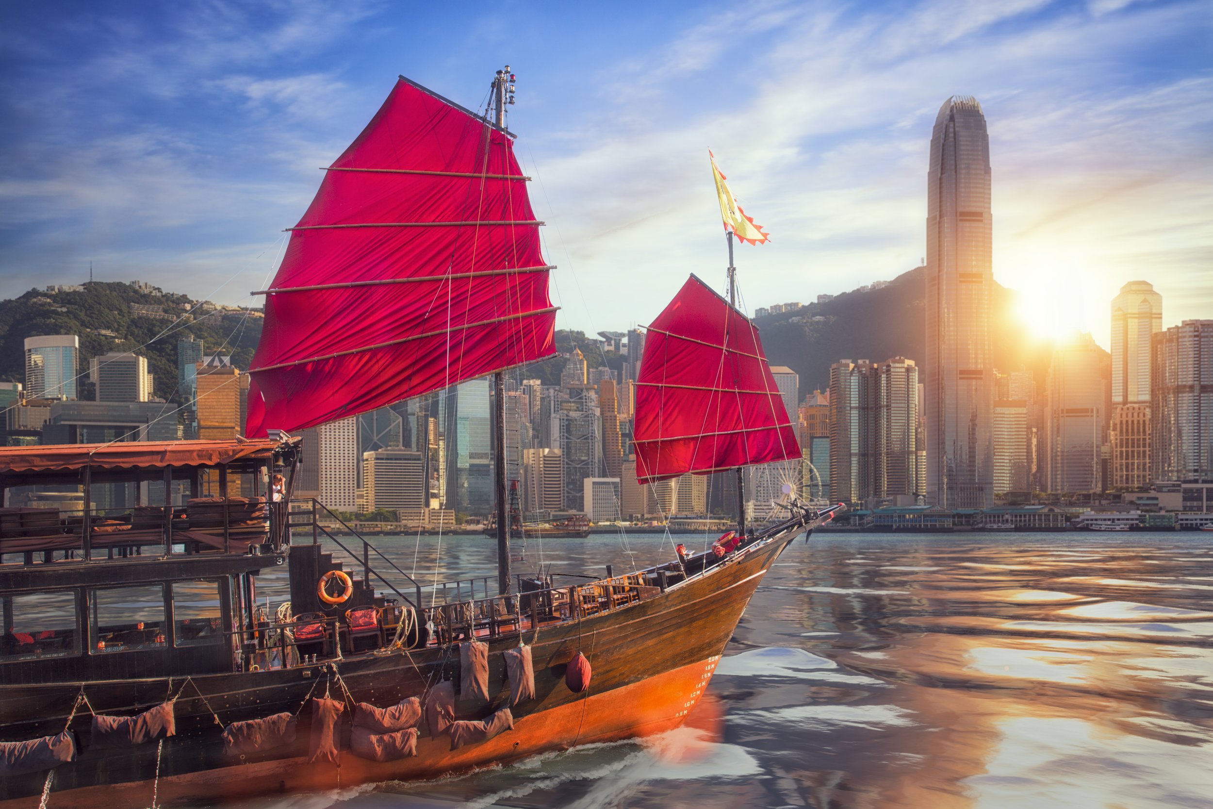 A traditional wooden sailboat with bright red sails on the water, with a city skyline and tall skyscrapers in the background during sunset.