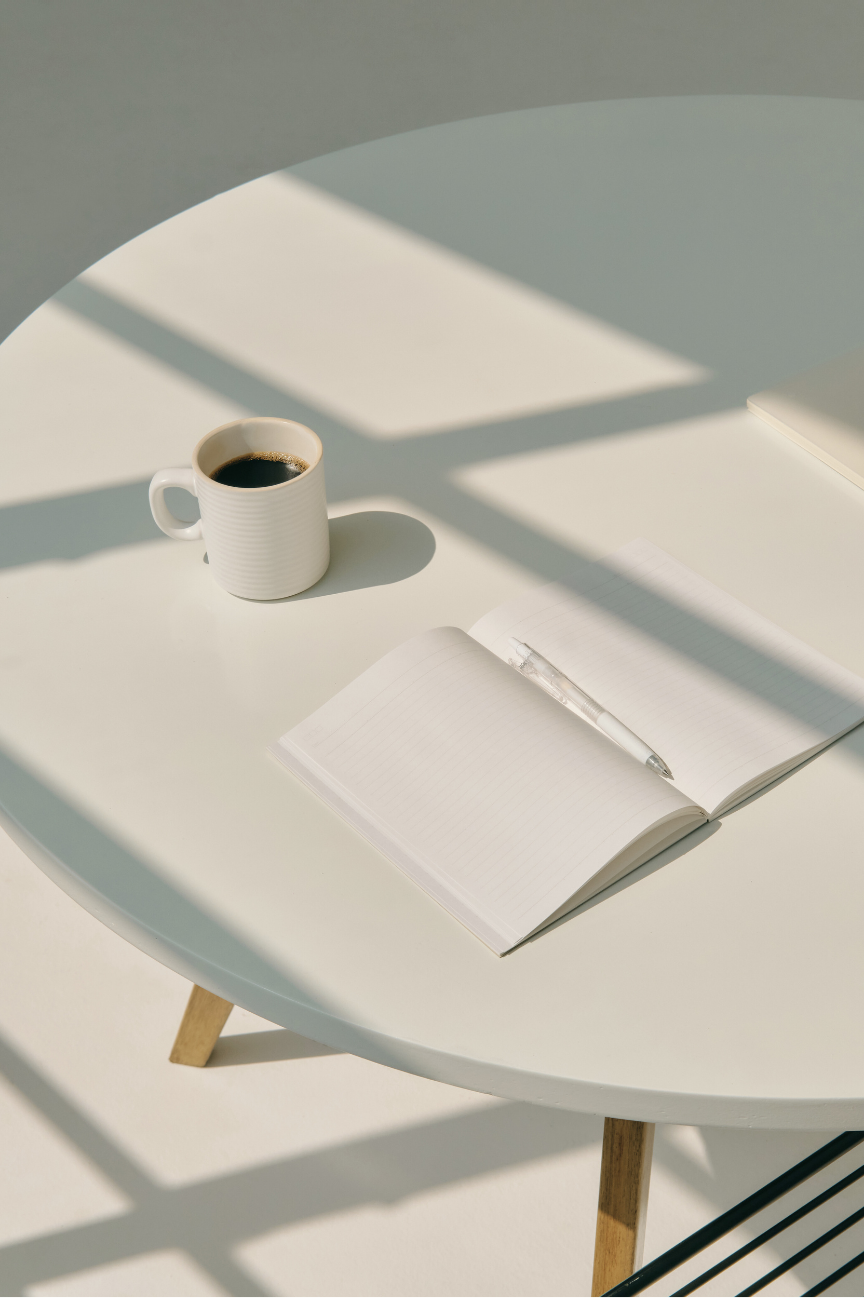 A white round table with a cup of black coffee, an open notebook with a pen on it, and a closed notebook. Sunlight creates geometric shadows on the table and the surface.