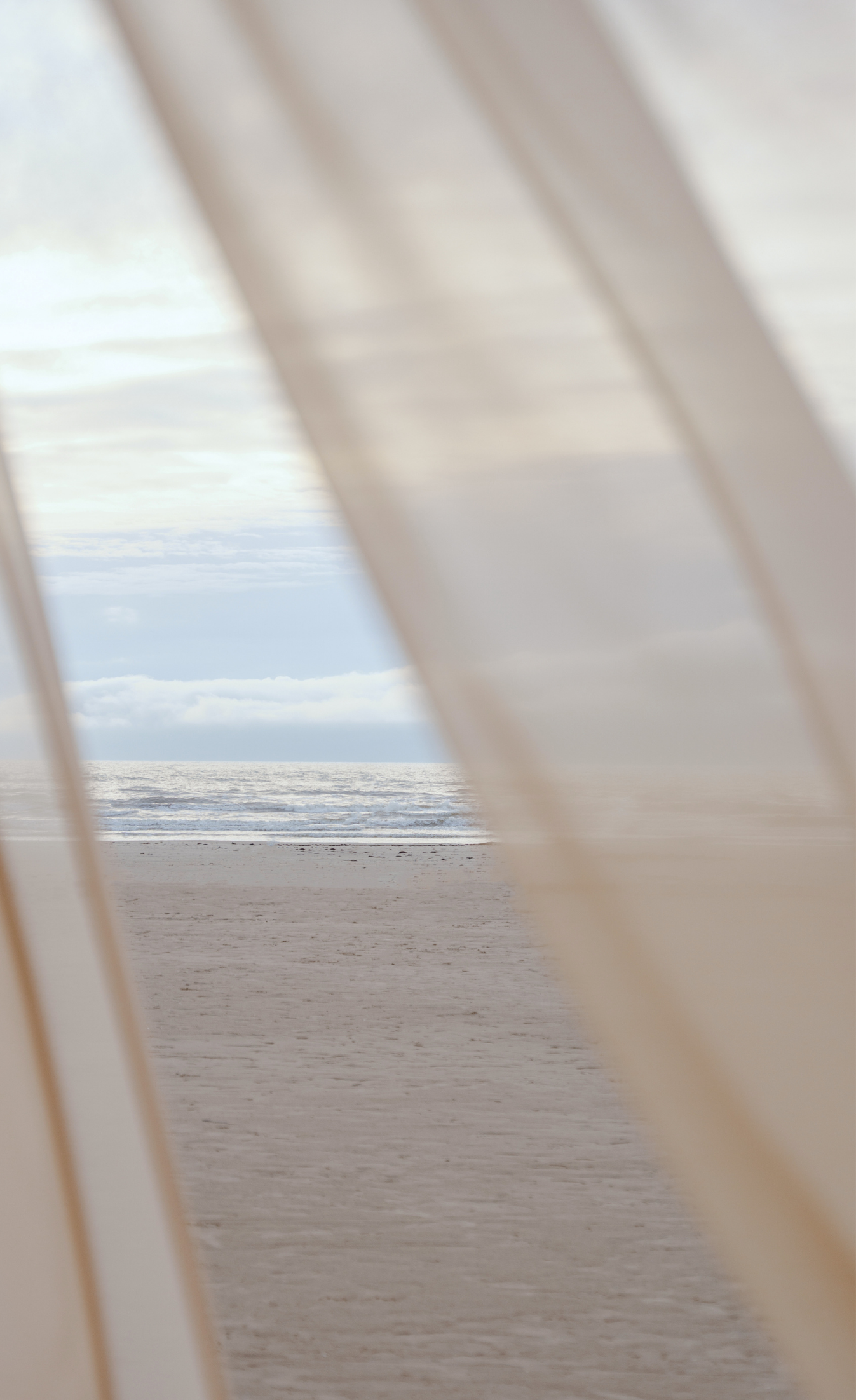 View of a sandy beach and ocean seen through white panels or curtains.