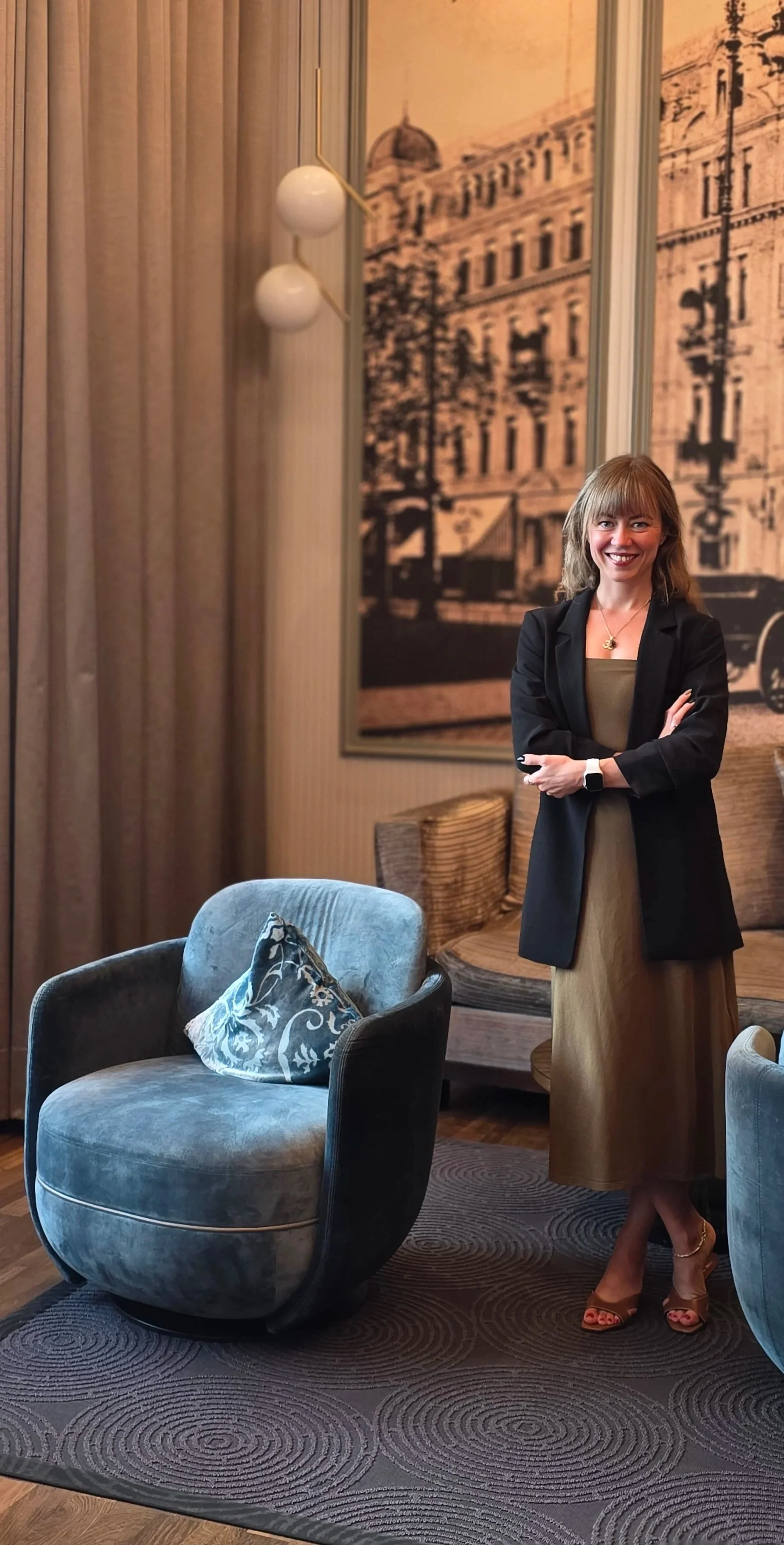 Frau im Business-Outfit steht lächelnd in einem eleganten Raum mit Pastellfarben, einer großen Schwarz-Weiß-Stadt-Ansicht an der Wand, einem bequemen grauen Sessel mit dekorativer Kissen und einem runden Holz-Tisch.