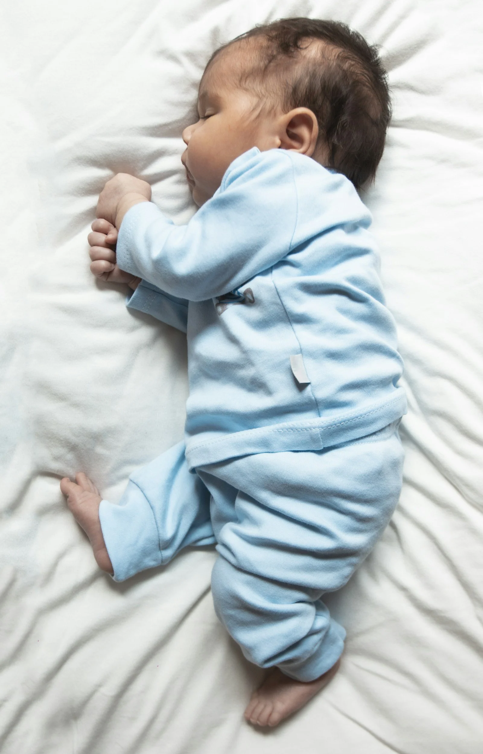 A sleeping baby lying on a white bed, wearing a blue outfit, with hands folded near the face and head turned to the side.