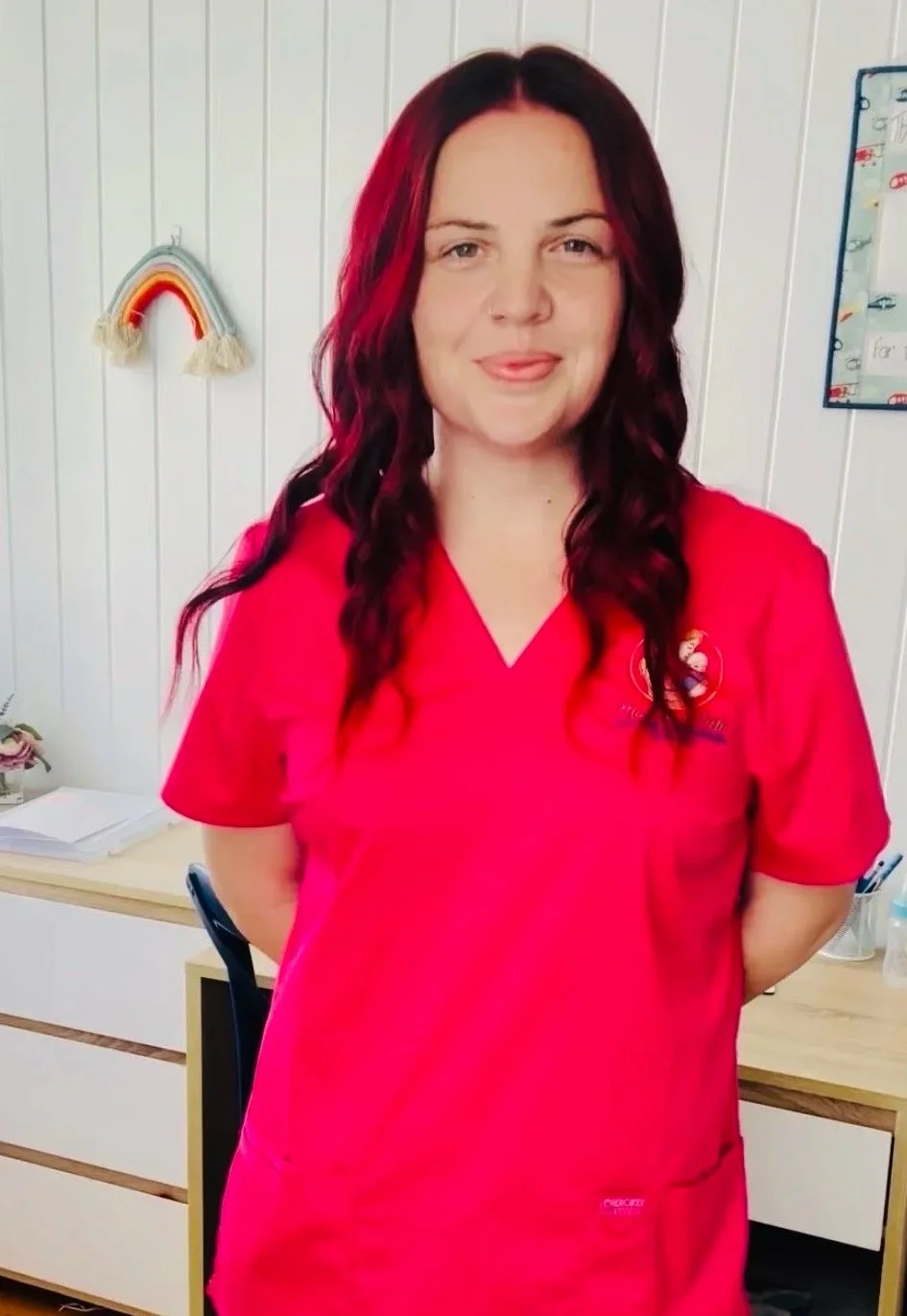 Woman with shoulder-length red hair standing in an office wearing a red shirt with a logo. Behind her are white wooden panel walls, a colorful rainbow wall hanging, and a framed whiteboard with writing and drawings.