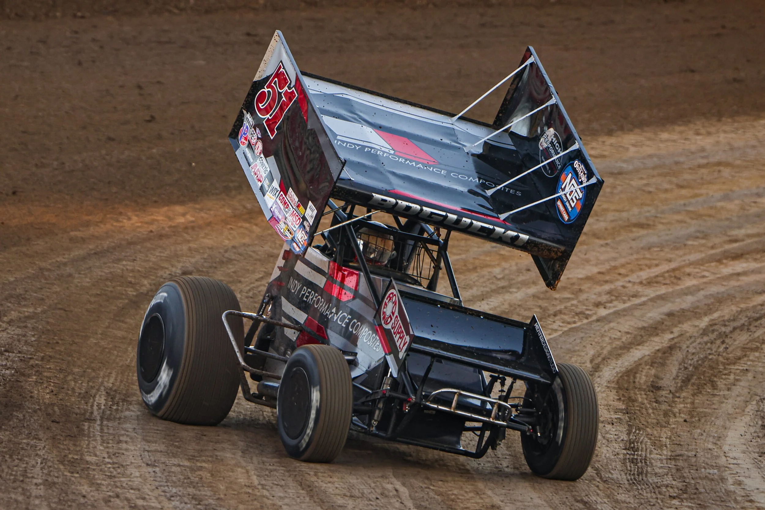 A black sprint car with advertisements and sponsor logos, including 'Lucas Oil' and 'INDY PERFORMANCE,' racing on a dirt track, with the rear wing tilted up and dirt kicking up behind it.
