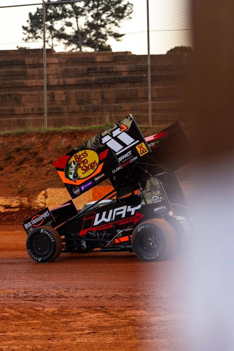 A black and red race car with a large rear wing, racing on a dirt track with a chain-link fence and trees in the background.