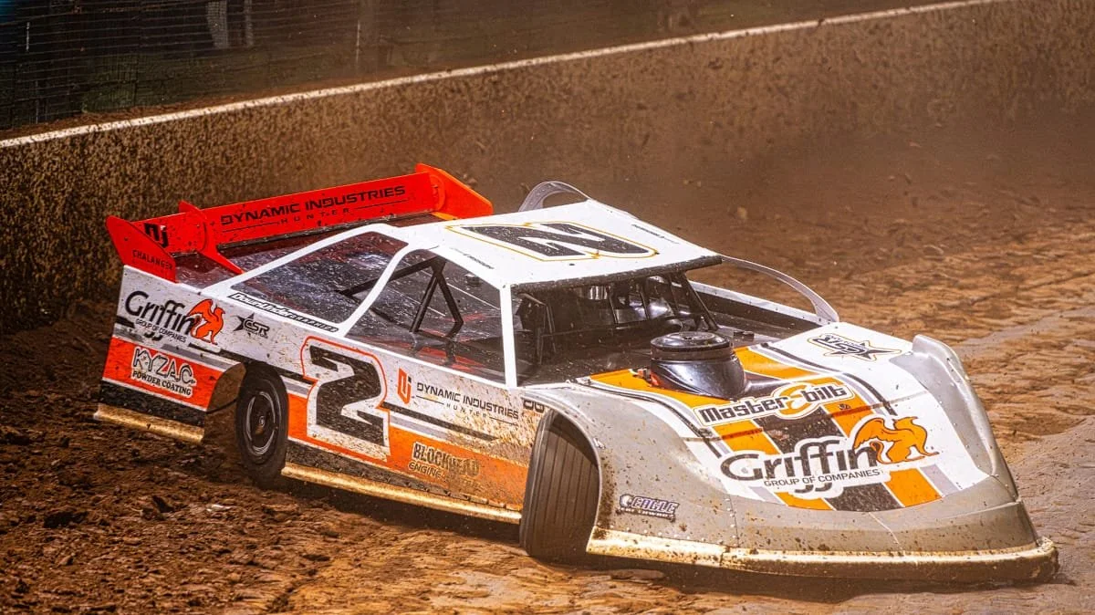 A white, orange, and black dirt track racing car with the number 2, kicking up dirt on a dirt race track.