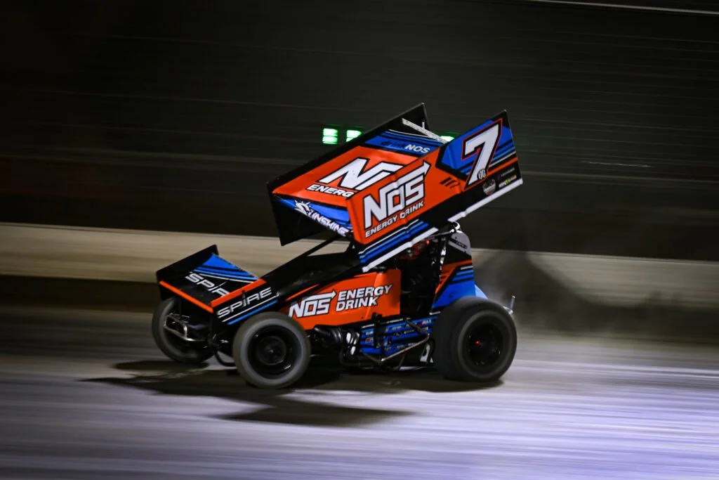 A sprint car racing on a dirt track at night, with a large wing on top displaying sponsorship logos.