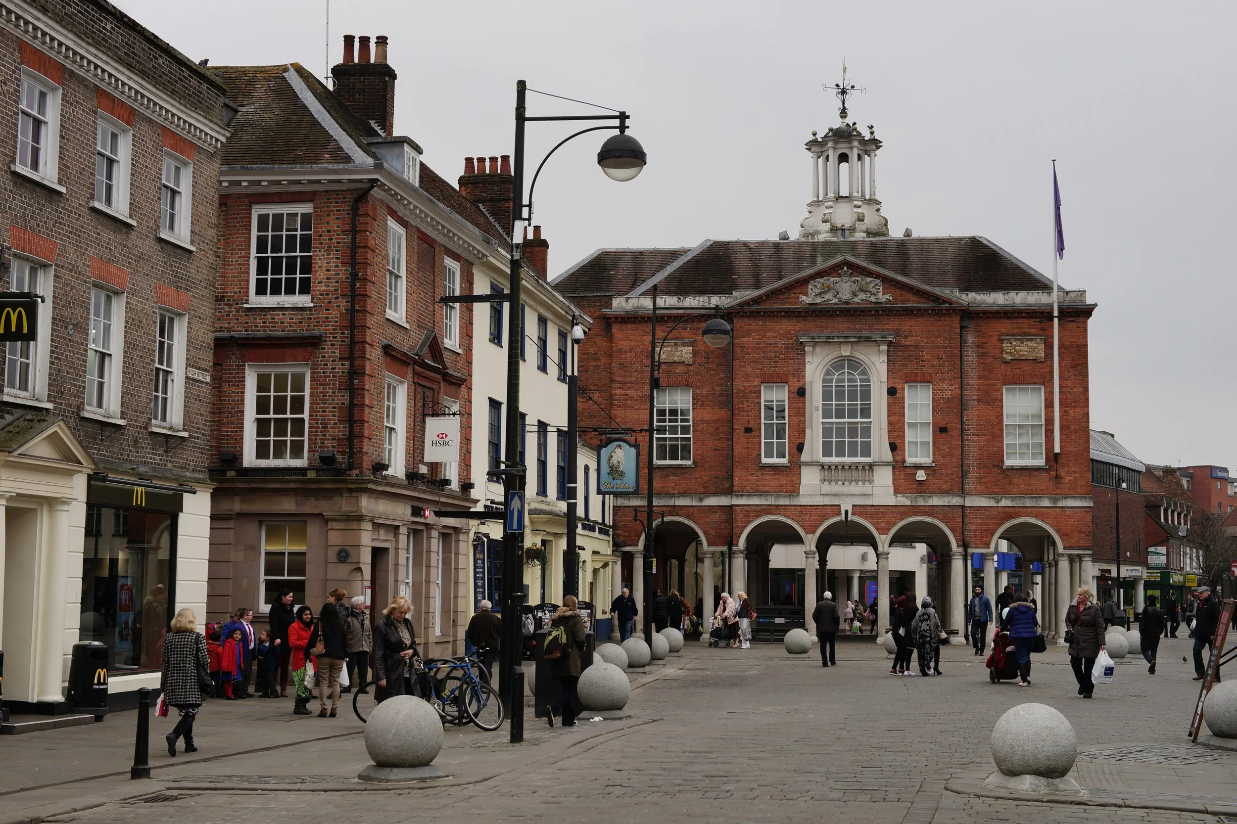 A busy town square with people walking and shopping, surrounded by historic brick buildings and a large red-brick building with a clock tower and decorative white accents. There are streetlights, benches, and spherical stone bollards along the sidewalk.