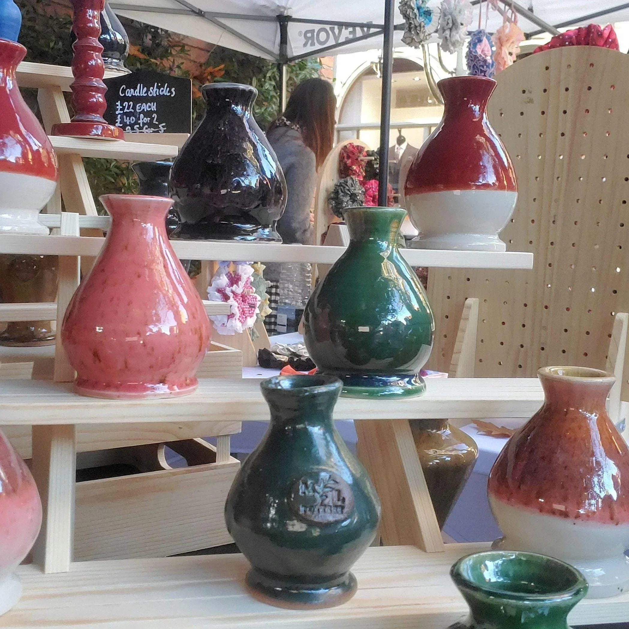 Display of various colorful ceramic vases and candle sticks at an outdoor market stand, with a person browsing in the background.