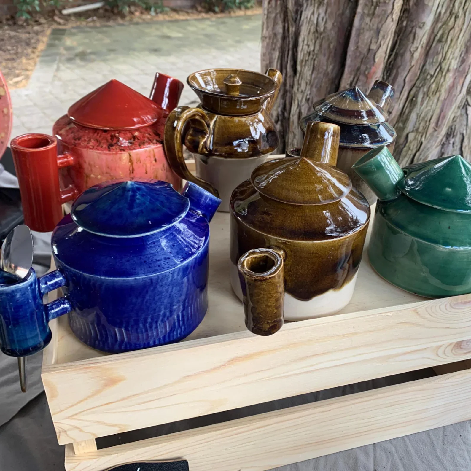 Colorful glazed ceramic teapots and pitchers with lids in red, brown, blue, and green placed on a wooden table outdoors near a tree.