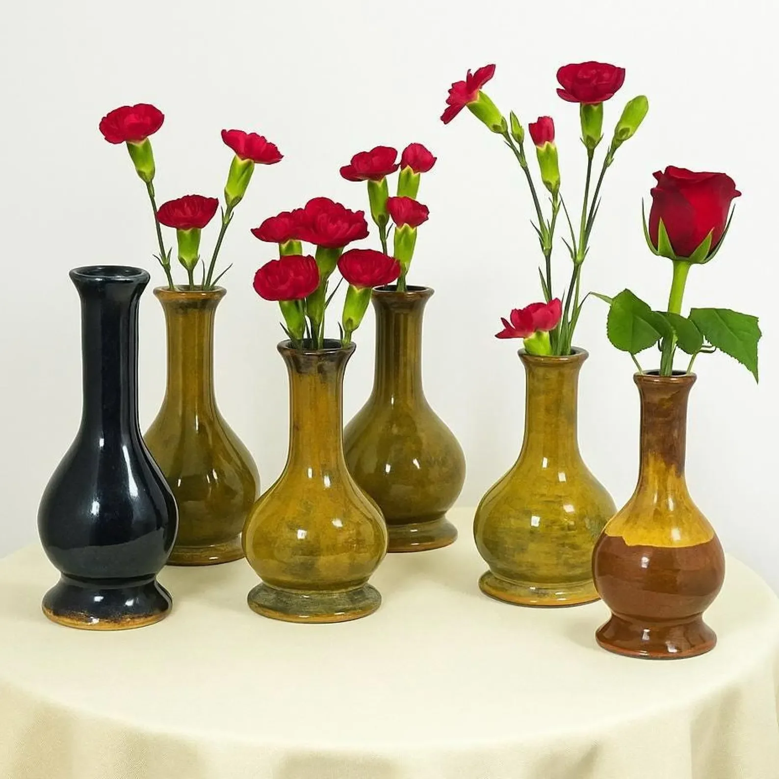 Seven ceramic vases in black and amber colors holding red carnations and rose flowers, arranged on a cream-colored surface against a plain white background.