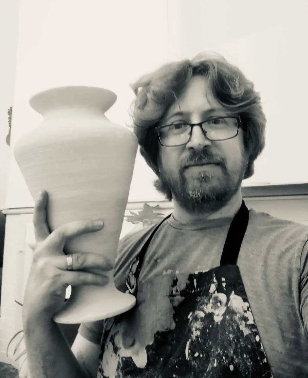 A man with glasses, curly hair, and a beard holding a large pottery vase in an art studio.