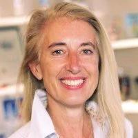 Frau mit blondem Haar und weißen Oberteil, lächelnd, im Hintergrund eine medizinische oder wissenschaftliche Umgebung