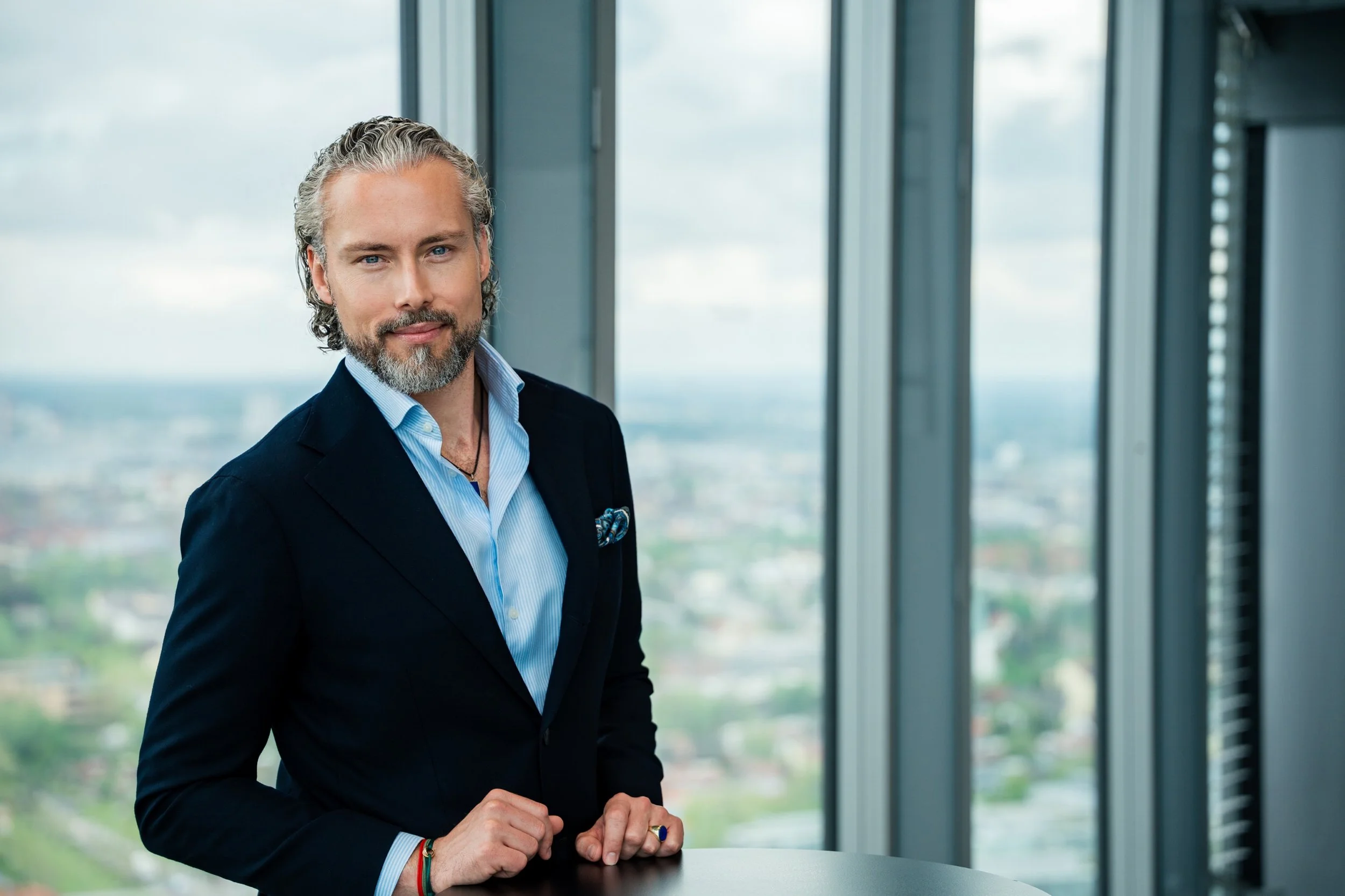 Ein Mann mit grauem Haar und Bart, im Anzug, steht in einem modernen Büro mit großen Fenstern und Blick auf die Stadt. Er blickt direkt in die Kamera.