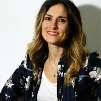 Eine Frau mit braunen, gewellten Haaren, trägt eine schwarze Jacke mit weißen Blumenmustern und ein weißes Top, lächelnd vor neutralem Hintergrund.