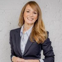 Ein Lächelndes Business-Porträt einer Frau mit langen, hellbraunen Haaren, in einem dunklen Blazer und einem blauen Hemd, vor einer grauen Wand.