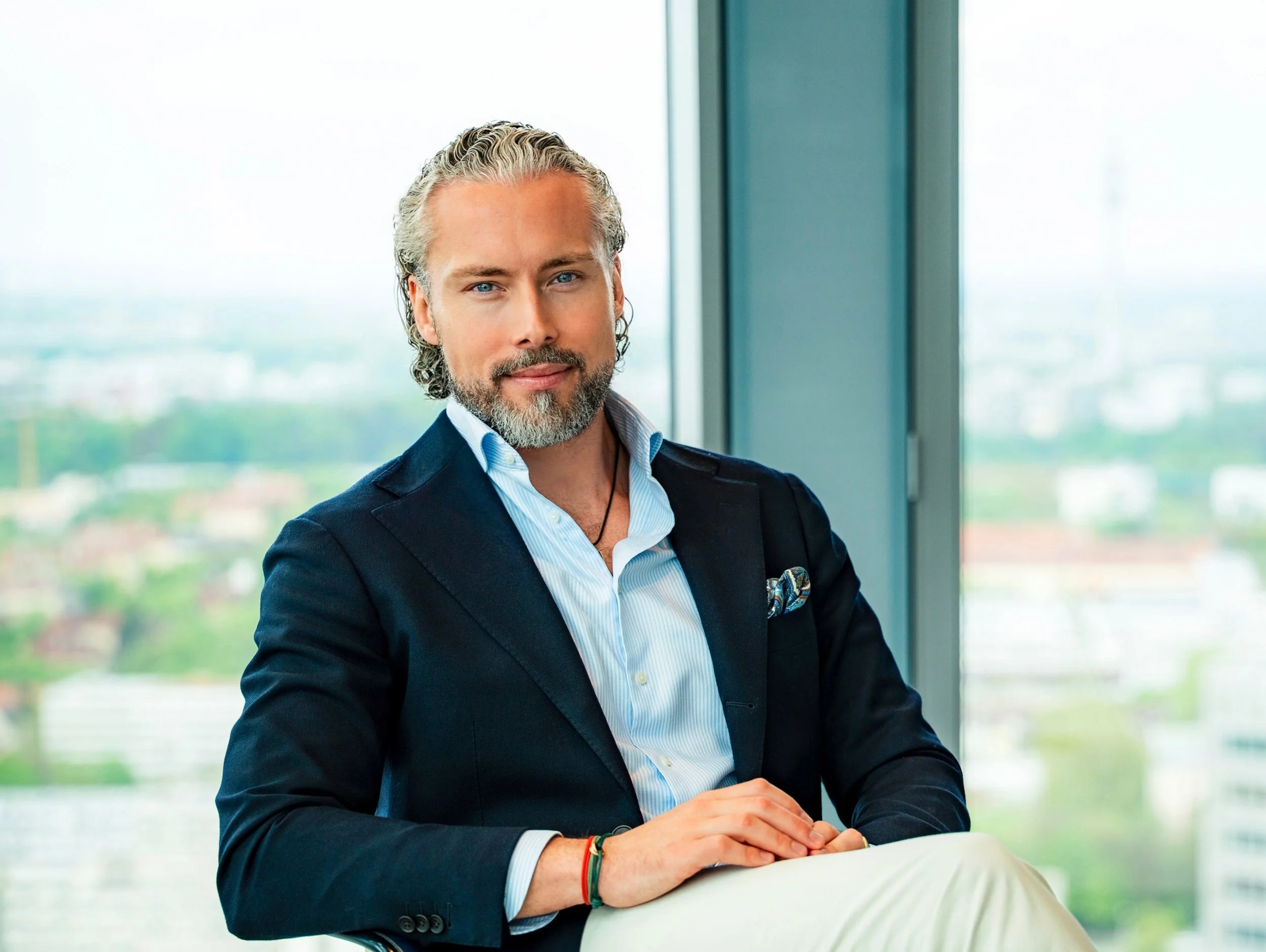 Dr. Moritz Behm mit grauem, welligem Haar und Bart, trägt einen blauen Anzug, sitzt vor einem großen Fenster mit Blick auf eine Stadt und lächelt leicht.