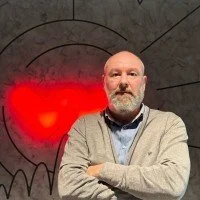 Mann mit Brille und grauem Pullover steht vor einer Wand mit rotem Herz und schwarzen Zeichnungen.