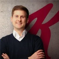 Ein Mann mit braunen Haaren, einem dunklen Pullover und weißem Hemd, der vor einem Hintergrund mit einem großen roten Logo steht.