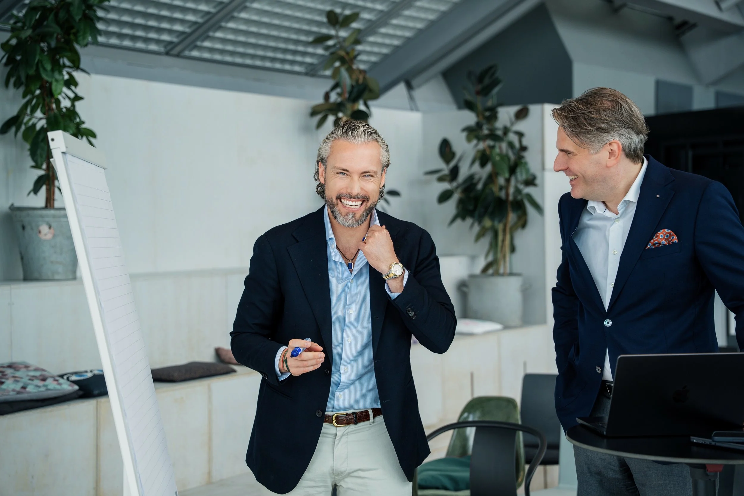 Zwei Männer in Anzügen lachen und sprechen in einem modernen, hellen Raum mit Pflanzen. Eine Person hält einen Marker und scheint eine Präsentation oder ein Meeting zu haben.