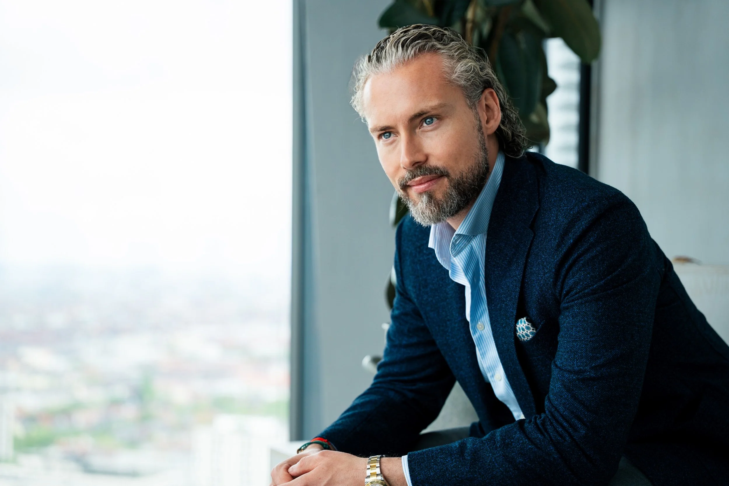 Ein Mann mit grauem Haar und Bart trägt einen blauen Anzug und sitzt an einem Tisch vor einem großen Fenster mit Stadtblick.