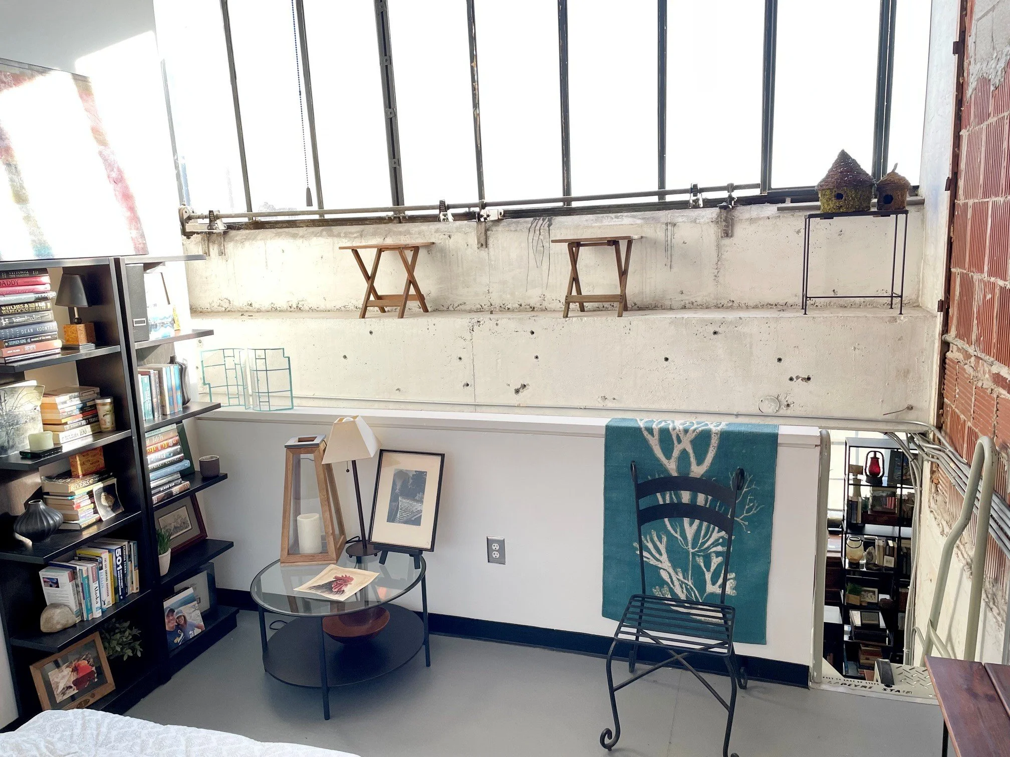 Interior of a loft-style apartment with large windows, exposed brick wall, bookshelf with books, framed photos, and decorative items, small sitting area with glass table, chairs, and artwork.