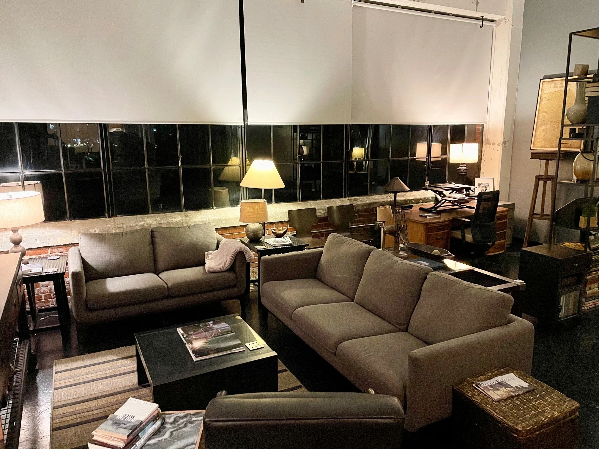 Living room with grey sofa, wooden coffee table, several lamps, a desk with chair, a shelf with vases, books, and decorative items, and large black-framed windows at night.