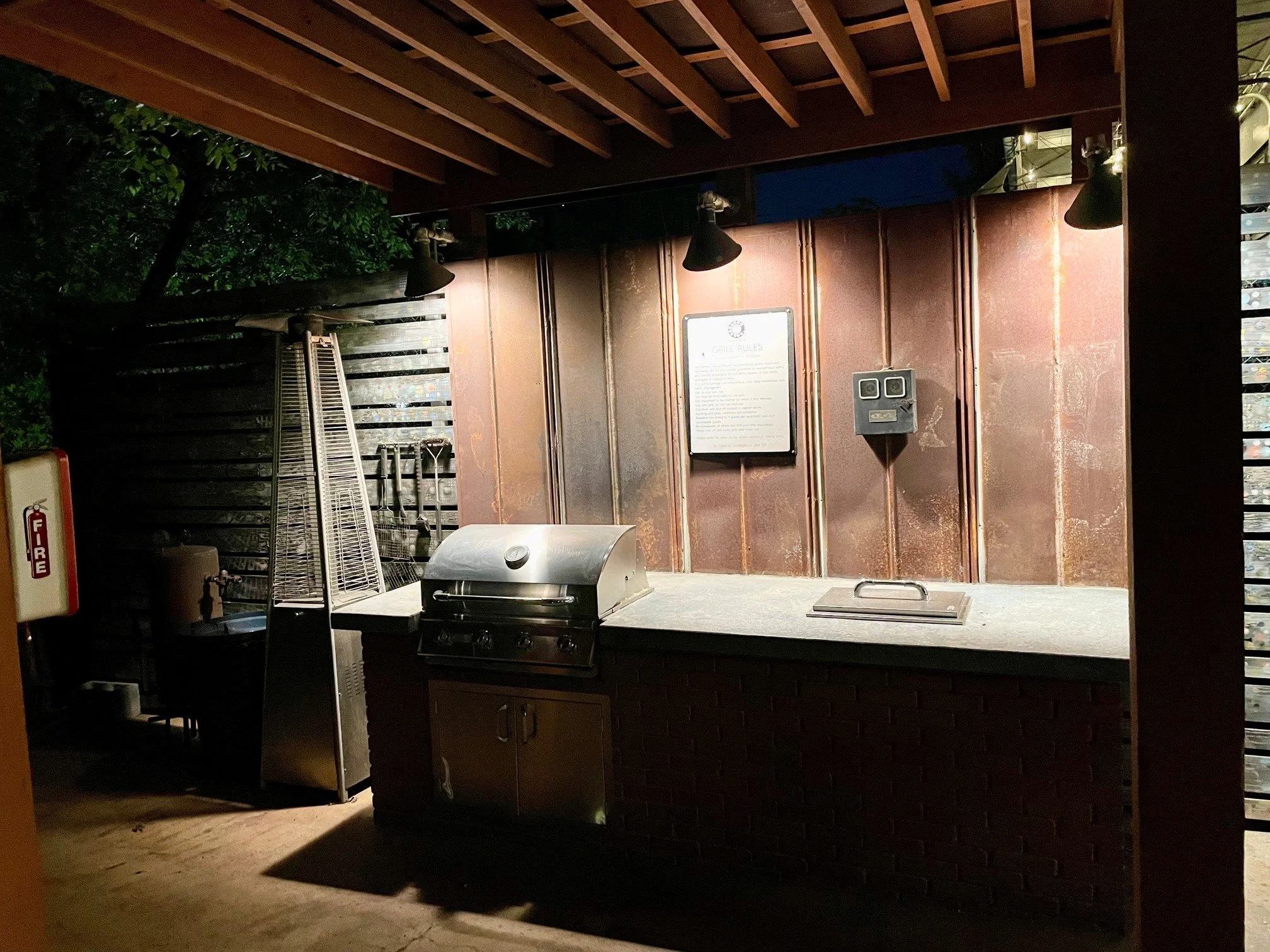 An outdoor barbecue area at night with a small grill, a window with a view of trees, and a sign with grill rules on a rusty metal wall, illuminated by three black wall lamps.