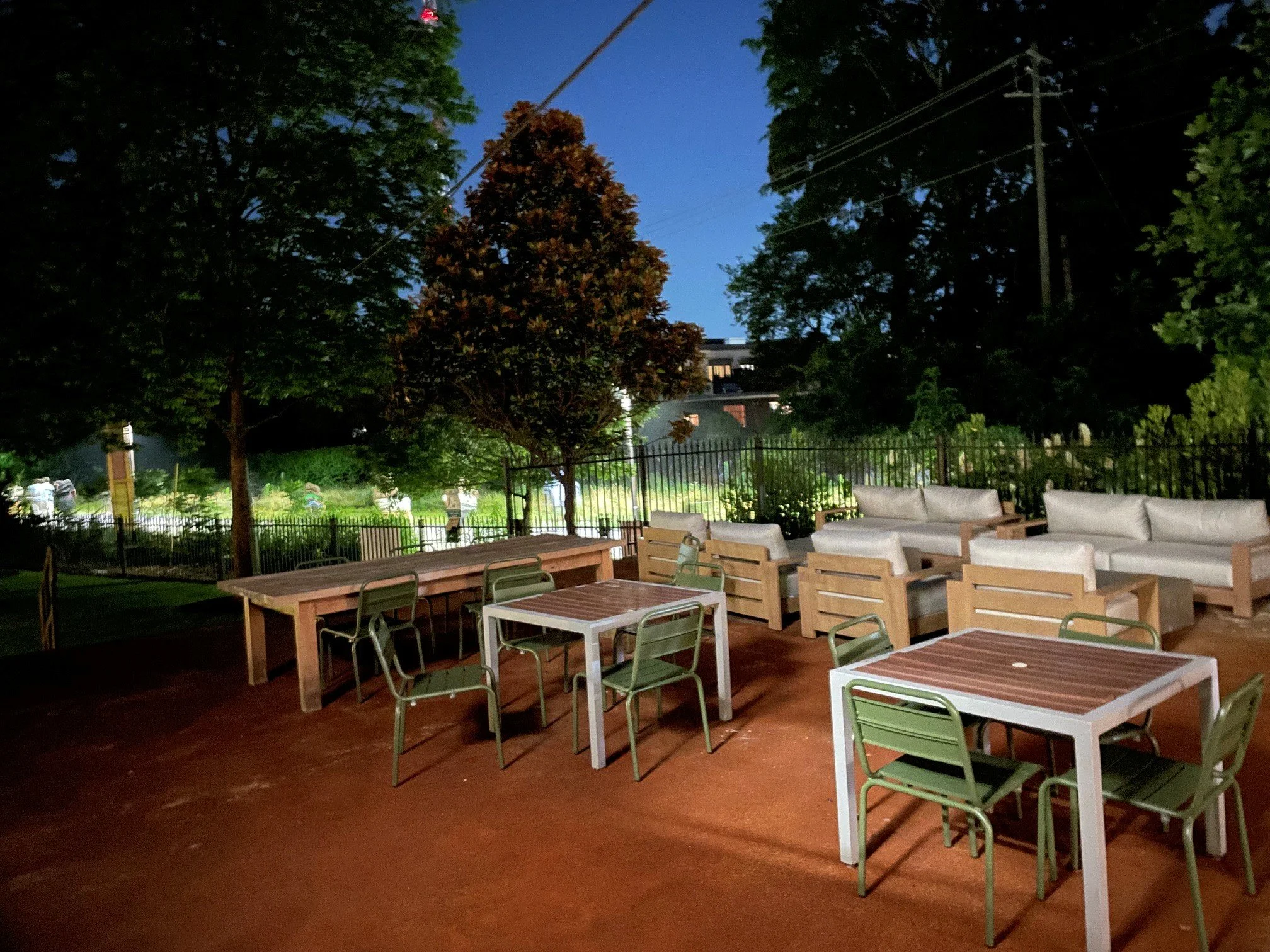 An outdoor patio area during dusk with wooden and metal tables, chairs, and white cushioned lounge seating, surrounded by trees and a black metal fence.