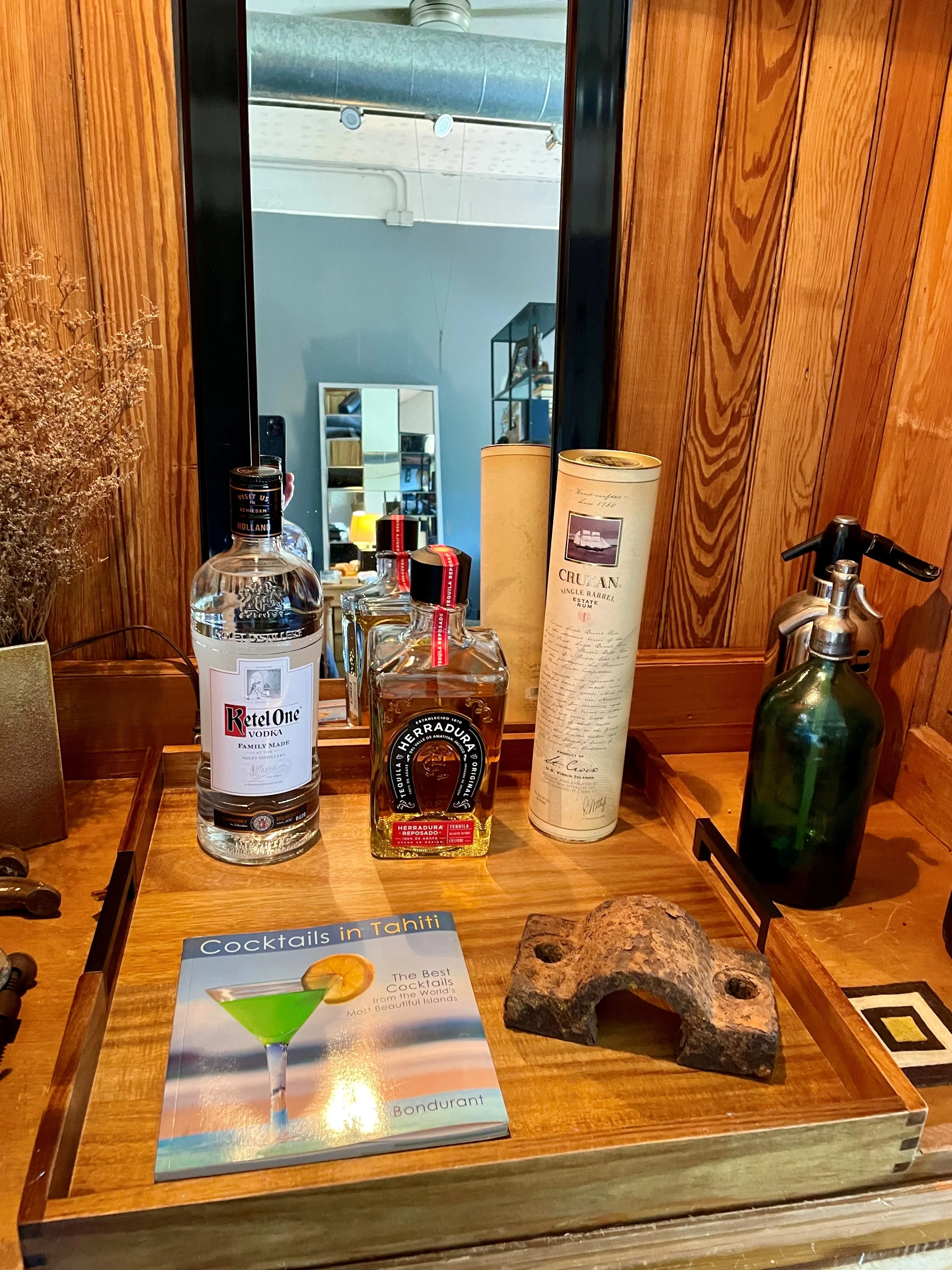 A wooden table holds several bottles of alcohol, a drink menu titled 'Cocktails in Tahiti,' and two decorative stone sculptures. A mirror on the wall reflects a room with a mirror, shelves, and various objects.