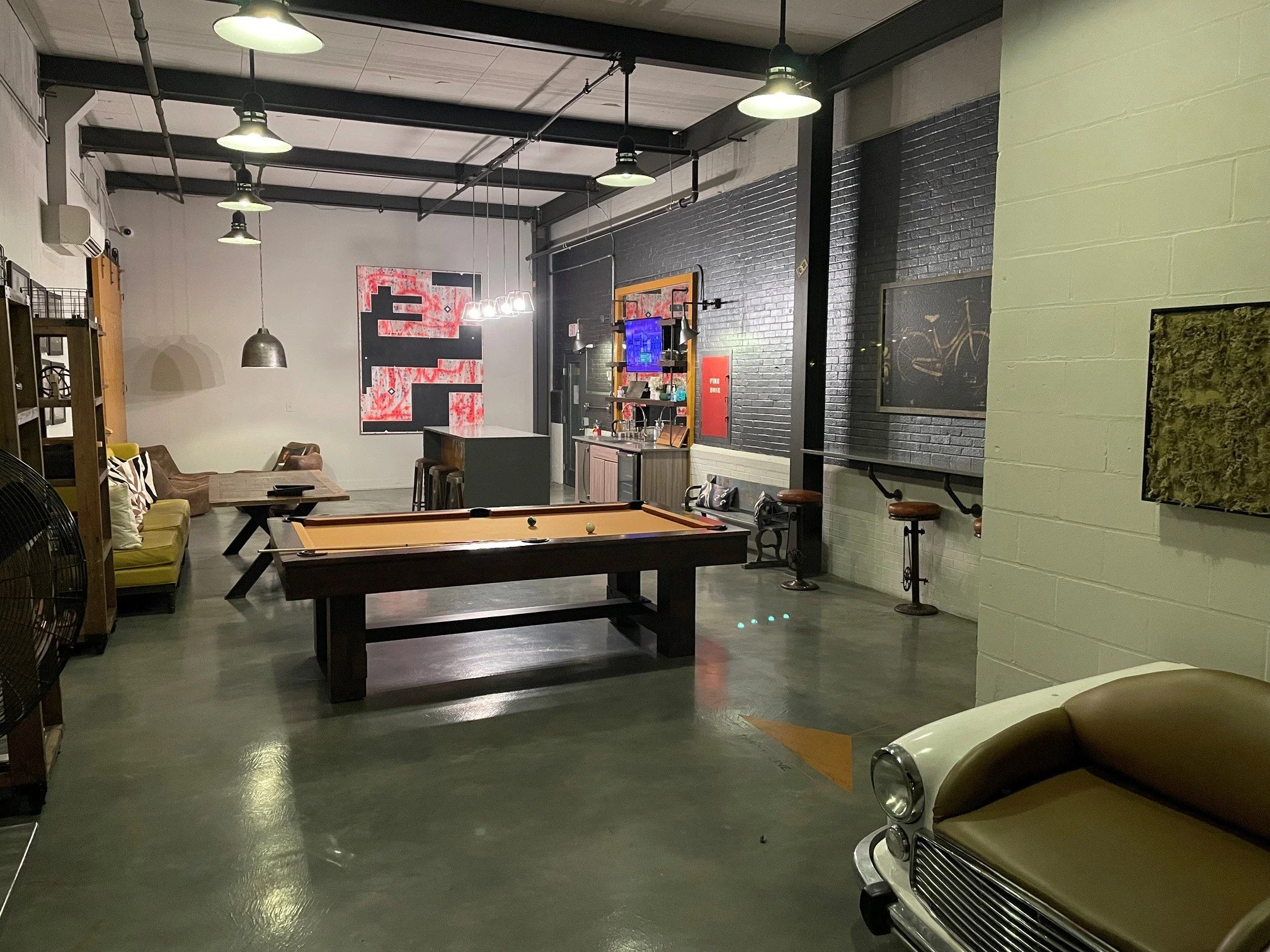An indoor recreational room with a pool table in the foreground, a vintage car at the right, a sofa and chairs on the left, artwork on the walls, a small kitchenette, and ceiling lights.