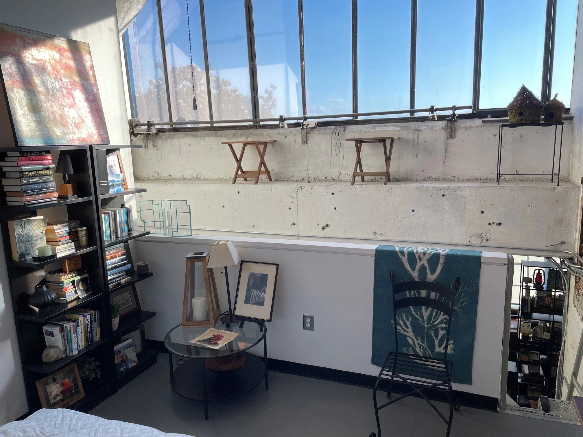 Living room with a bookshelf filled with books in the corner, a small glass-top table with framed photos and a magazine, an armchair with a patterned throw, a small side table with a framed photo, a window with a ledge and decorative birdhouses.